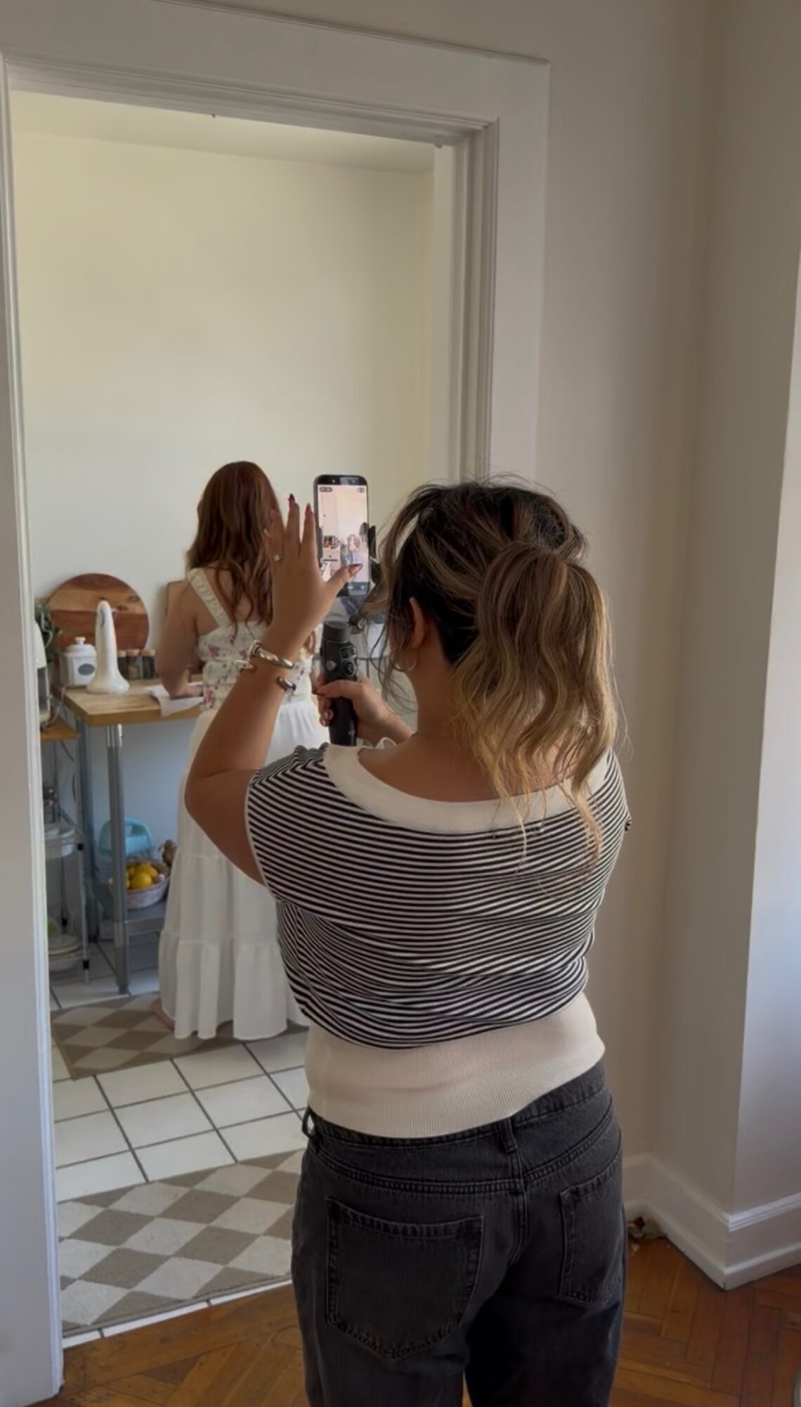 Person taking a mirror selfie in a room with a woman in the background, using a smartphone.