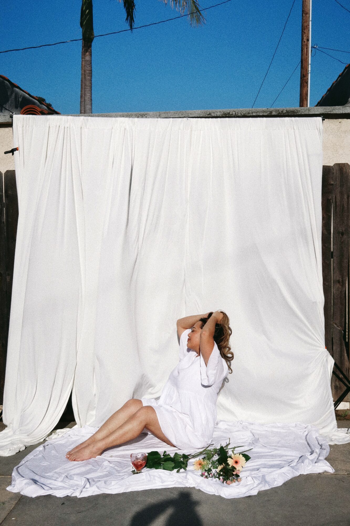 Person sitting on white fabric with flowers, head tilted back, in front of white curtain backdrop outdoors.