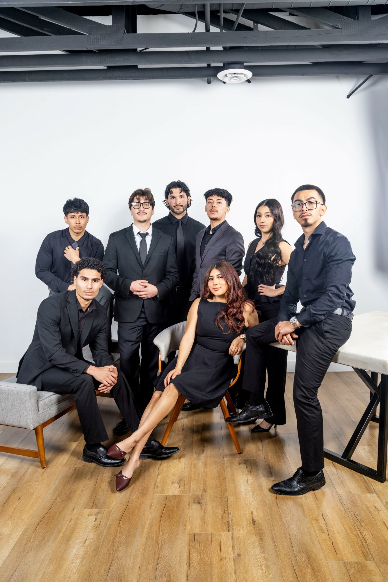 Group of eight professionally dressed people posing in a room with wooden floor and white wall.