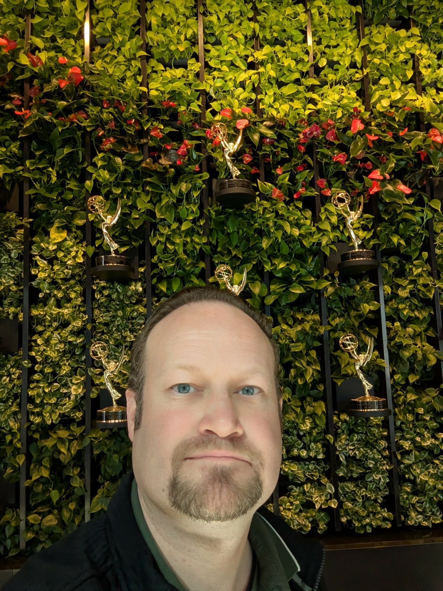 A man with short hair and a beard, wearing a dark jacket, stands in front of a lush green and flowering hedge.