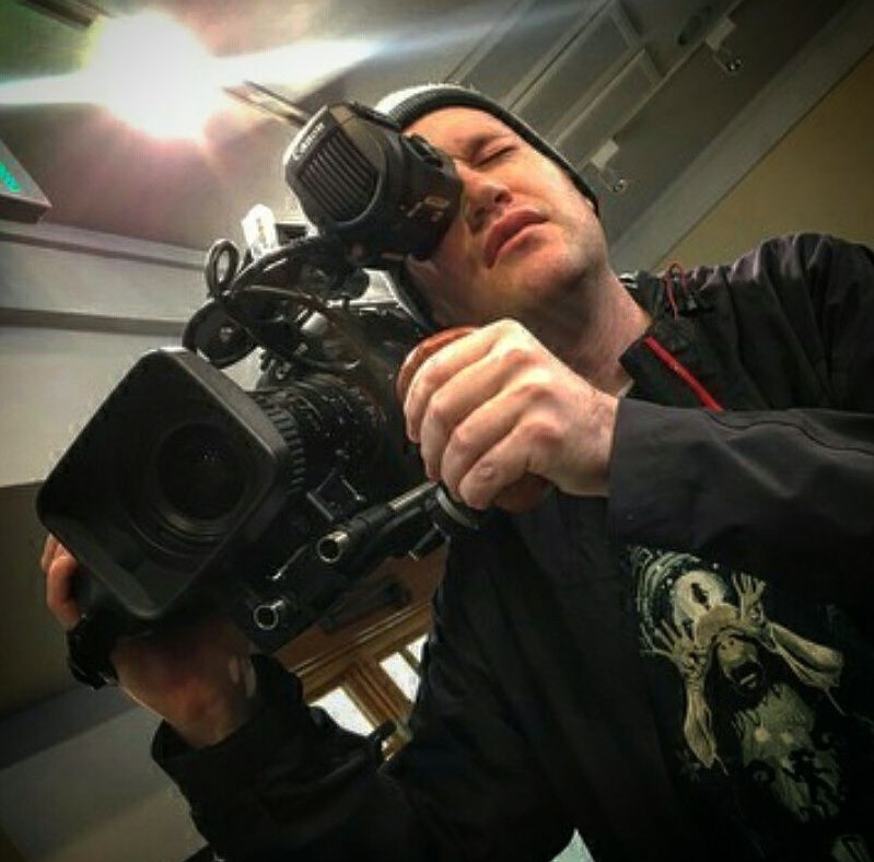 Person holding a professional video camera, looking through the viewfinder, indoors with ceiling lights visible.