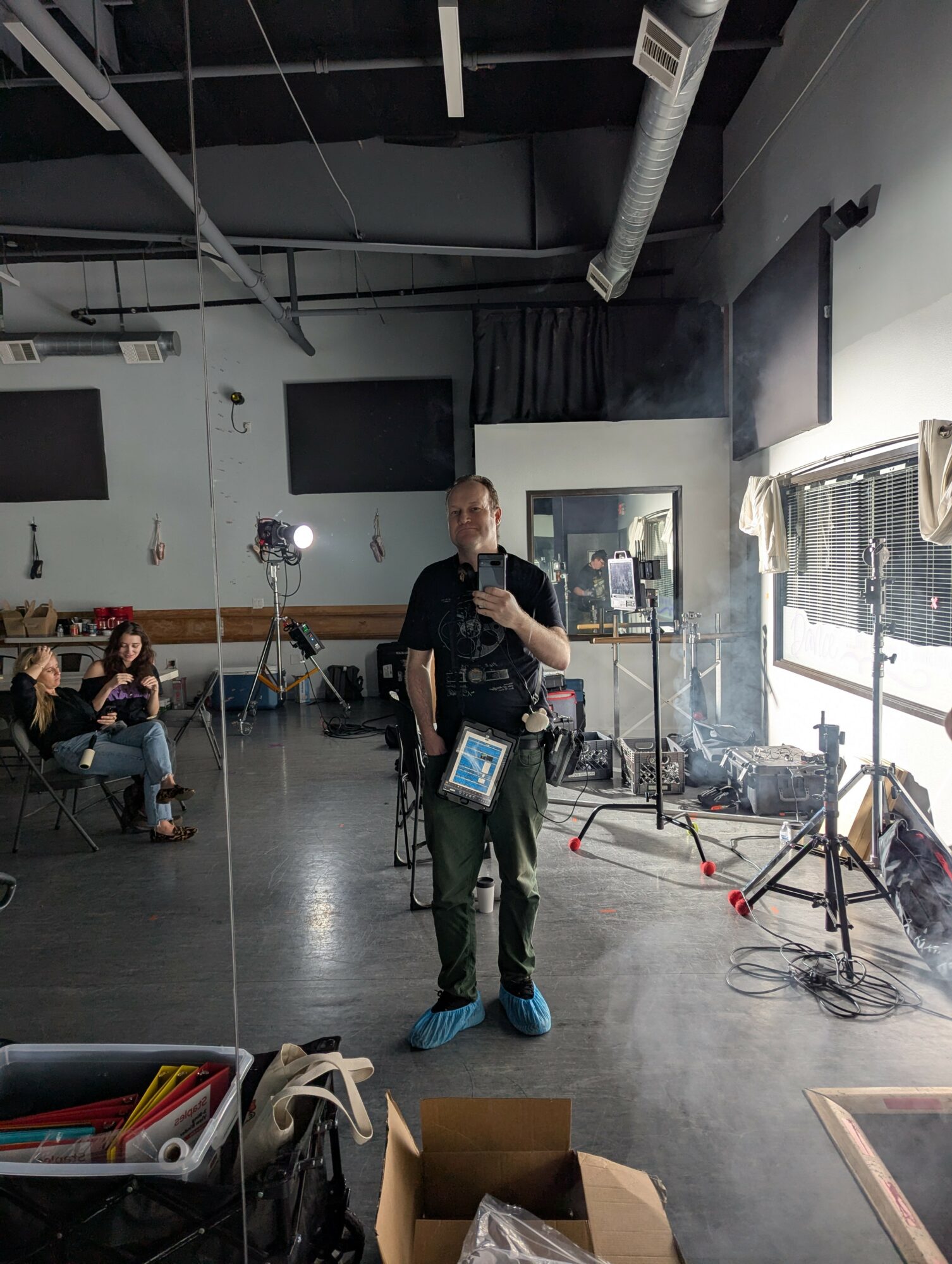 Person taking a selfie in a room with equipment, people sitting, and large windows with blinds.