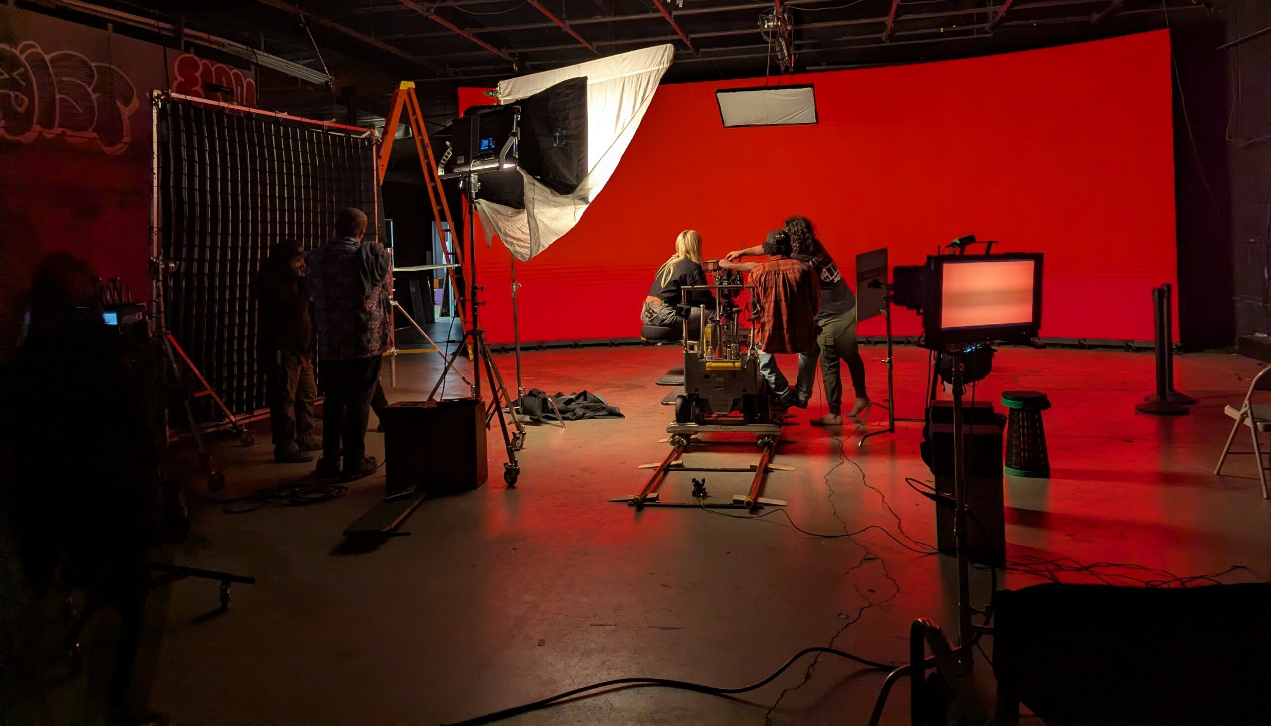 Film studio with crew preparing equipment and camera setup, red wall background, dim lighting, and various equipment scattered.