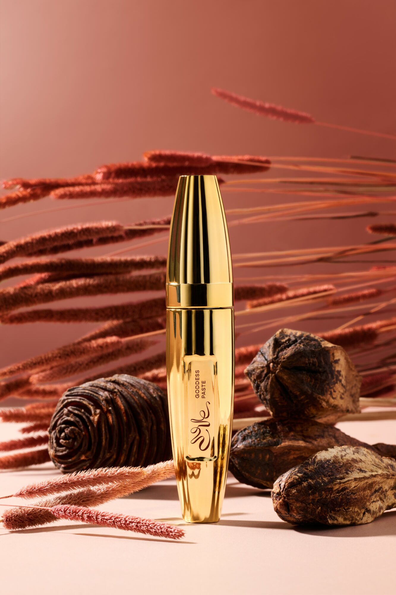 Gold-colored cosmetic bottle stands among rocks and pink decorative elements against a pink background.