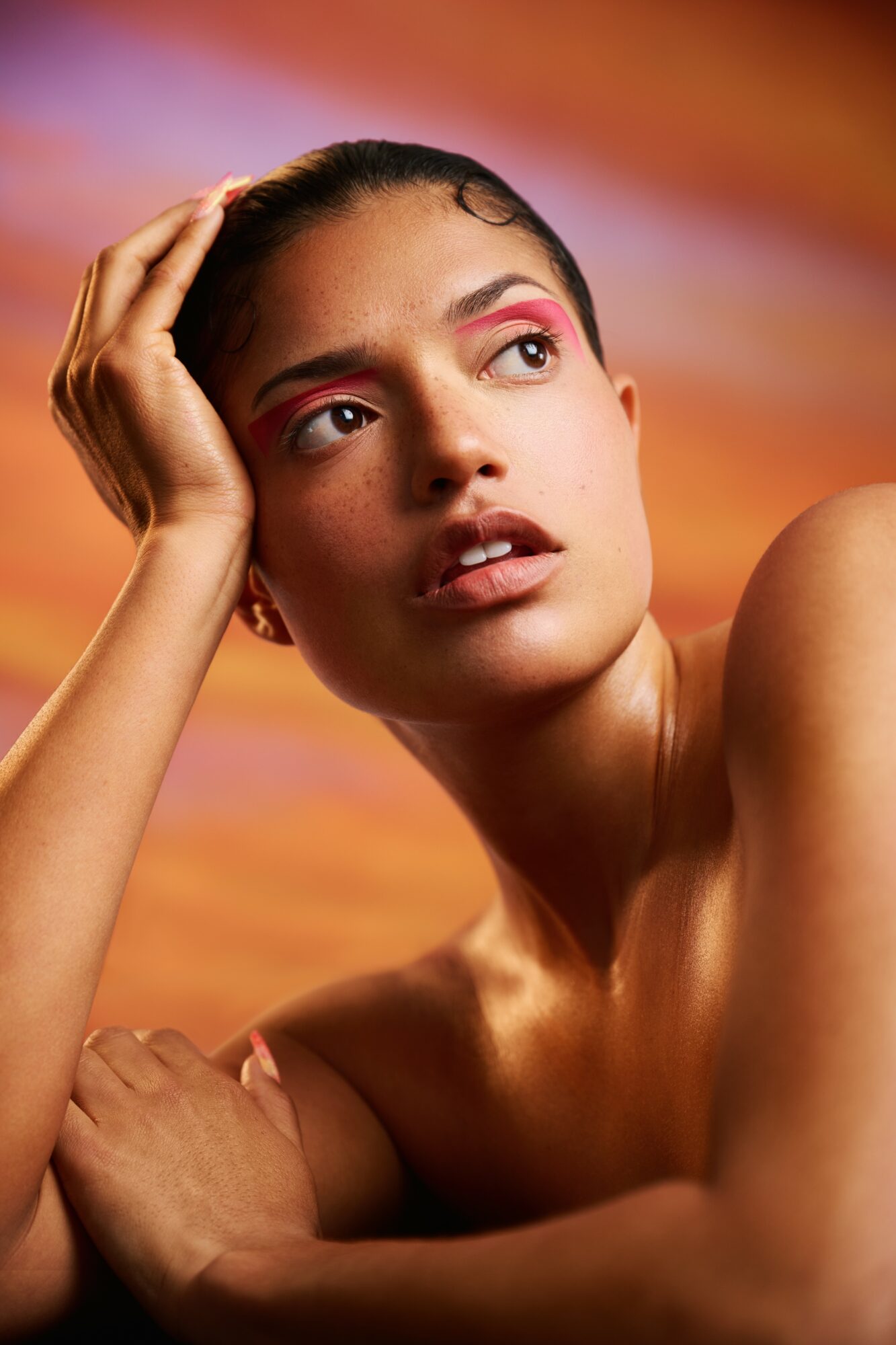 Young woman with pink eyeshadow resting her head on her hand, looking away, with a colorful sunset background.