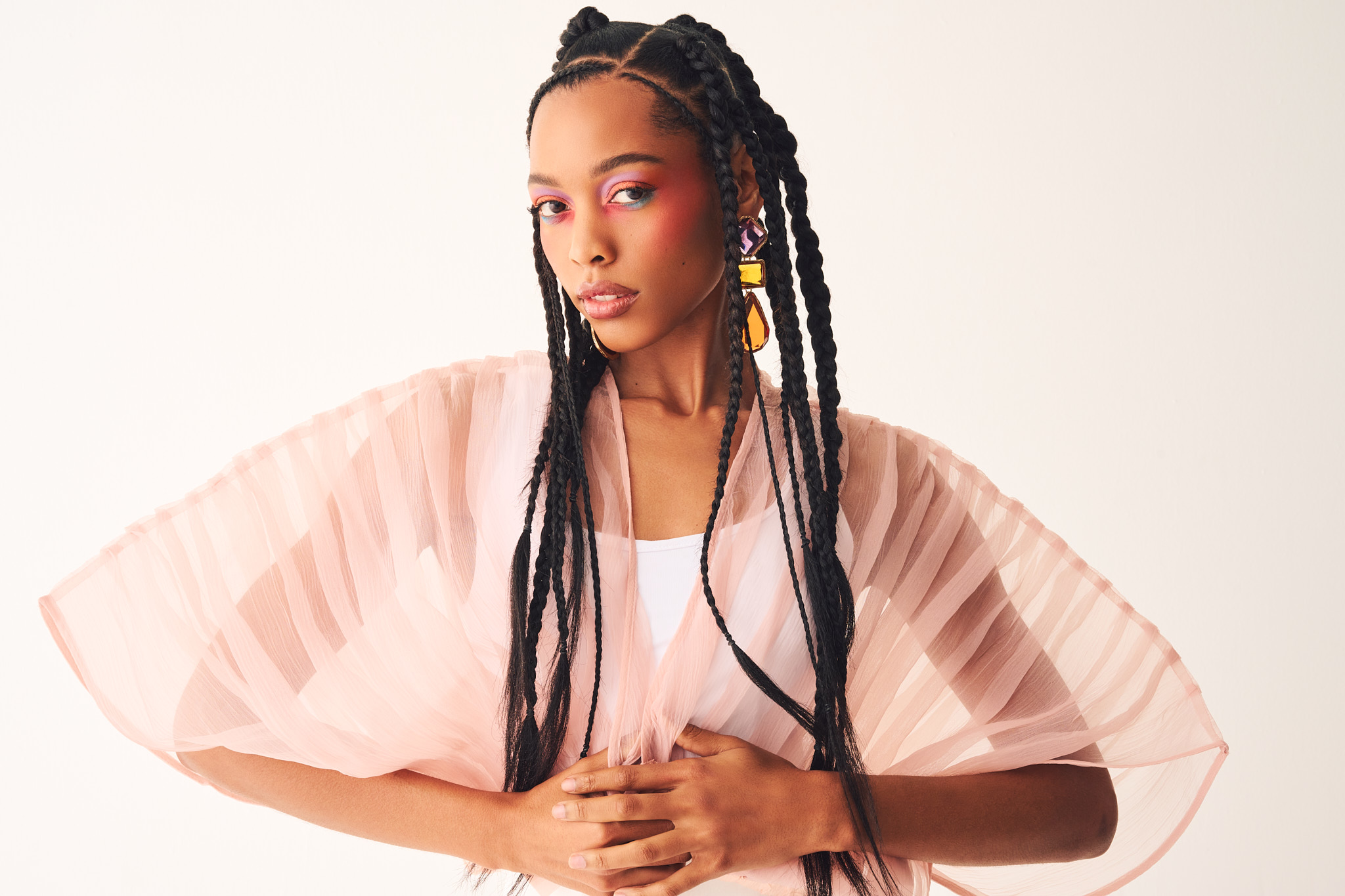 Young woman with long braided hair wearing a light pink pleated dress, standing against a plain background.