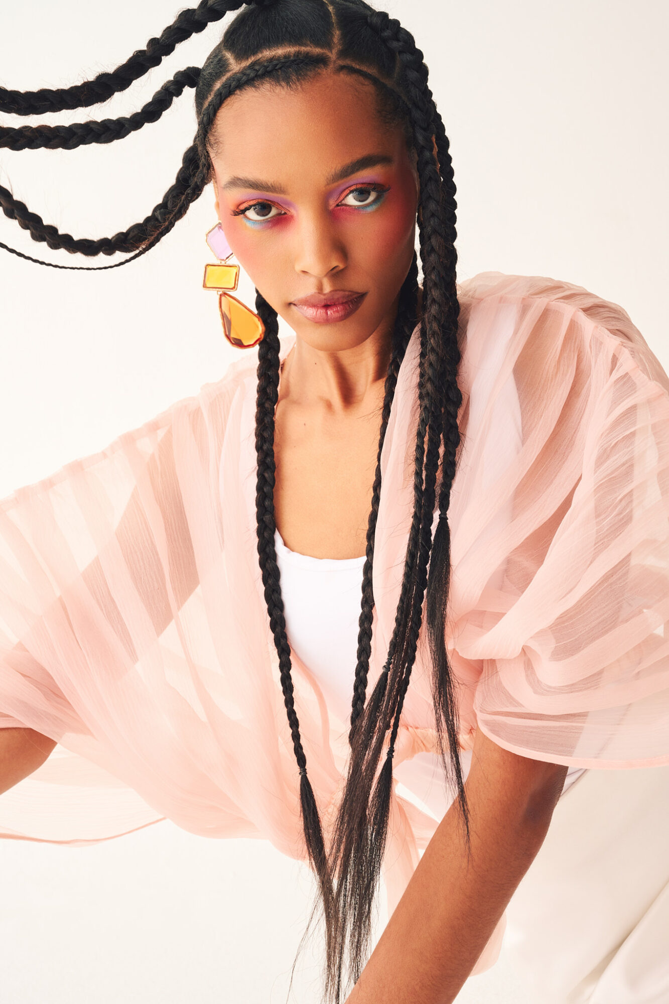 Young woman with braided hair, wearing colorful earrings and a light pink sheer top, posing against a plain background.