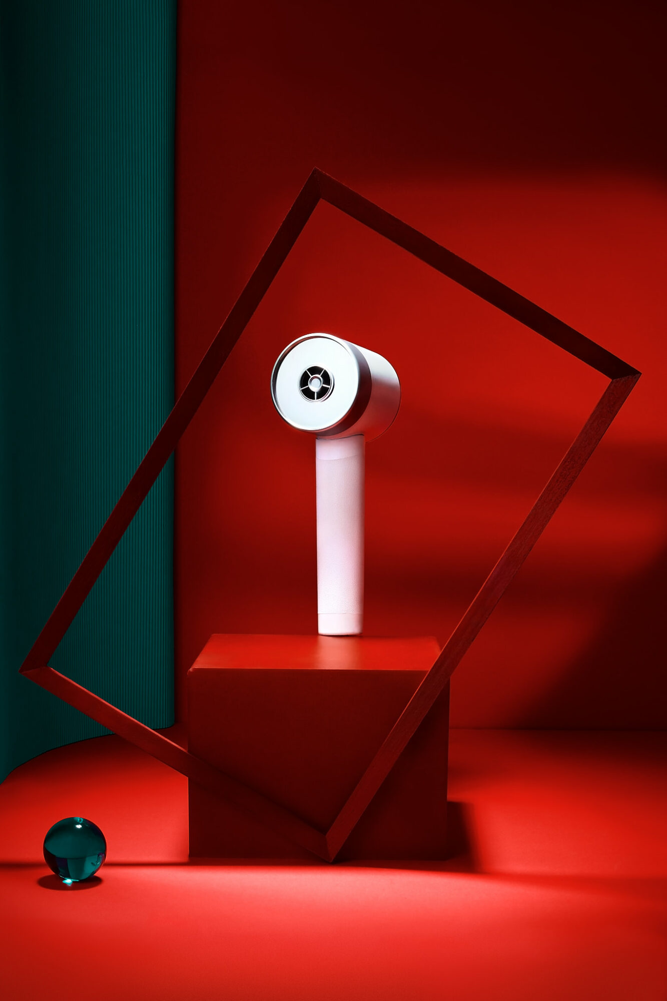 Object resembling a speaker on a white stand inside a tilted red frame, with a small black sphere nearby, against a red background.