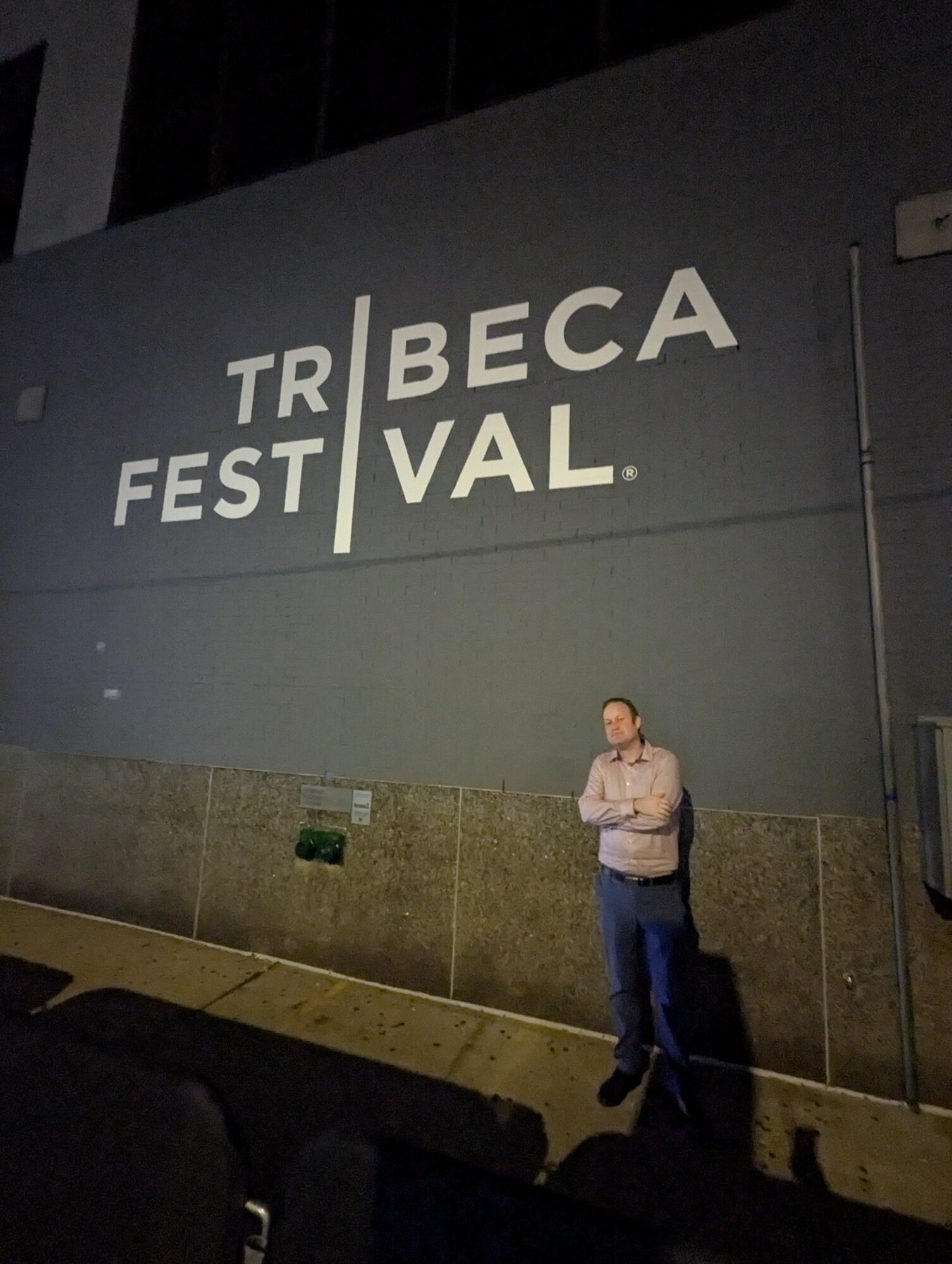 Person standing against a wall with festival signage, night setting, shadow cast on ground.