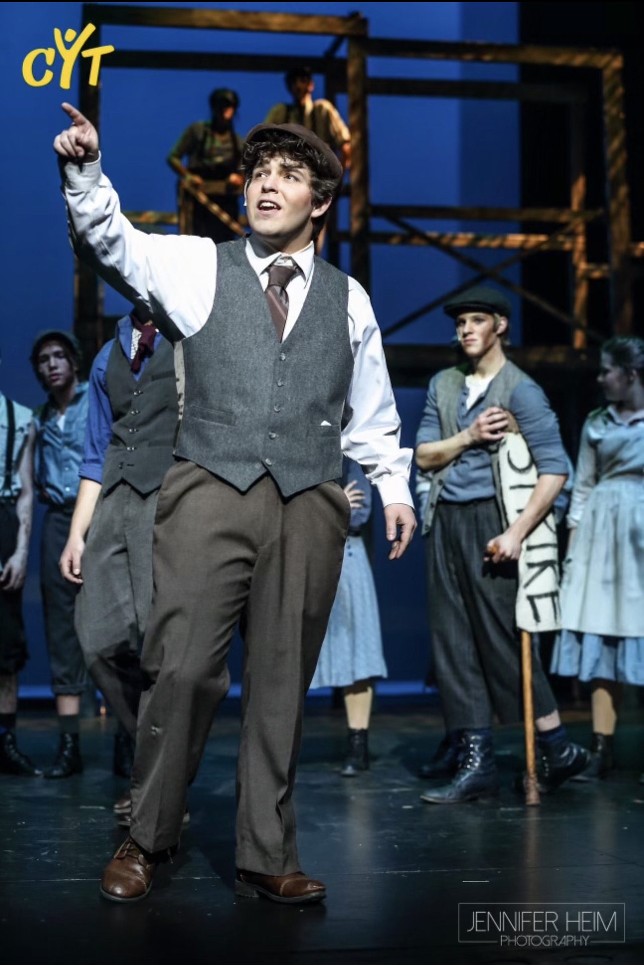 Young man in vintage clothing pointing upward on stage with other performers in background.