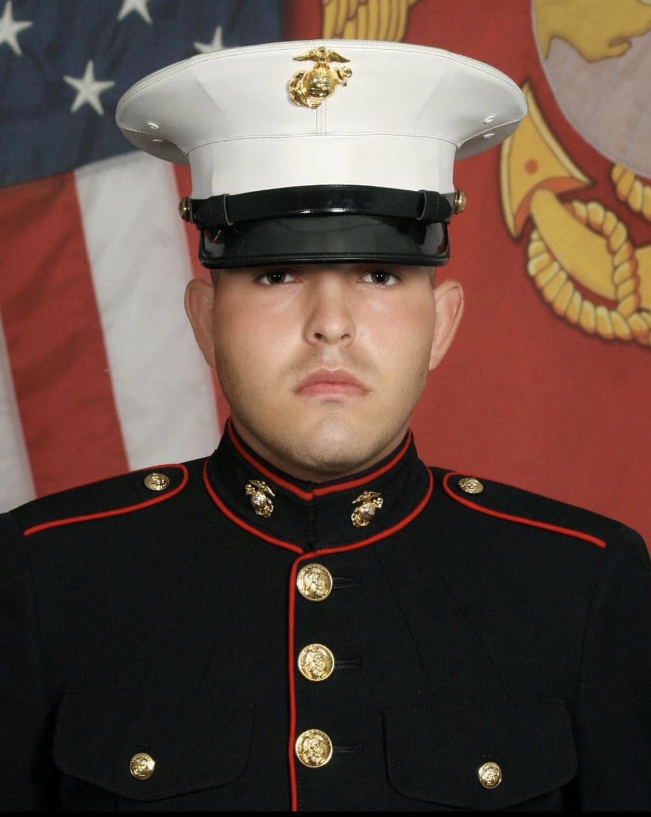 Young man in Marine dress uniform with white cap, black jacket, and gold buttons, standing in front of flags.