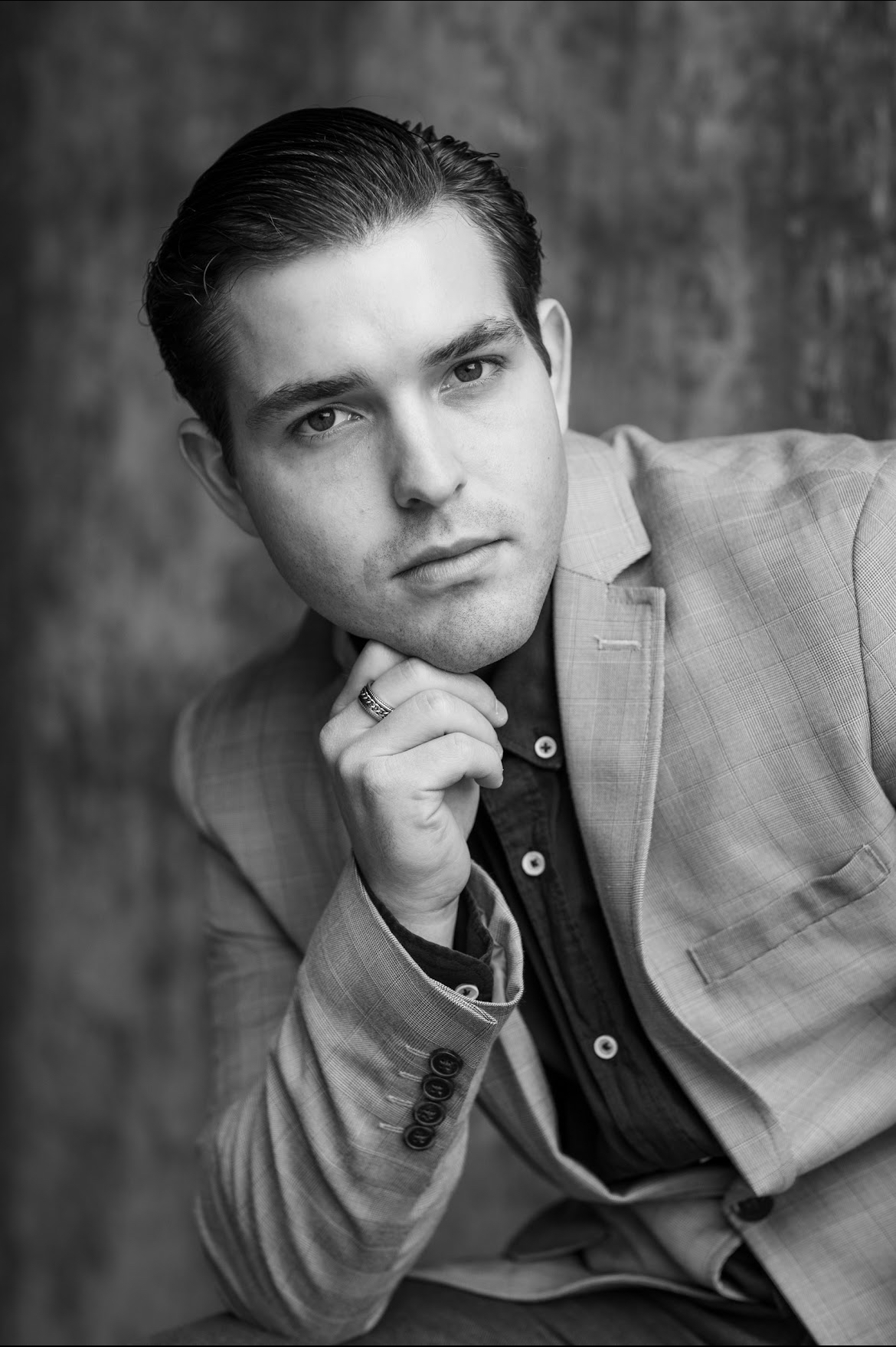 Young man with slicked-back hair, wearing a suit jacket and shirt, resting his chin on his hand.