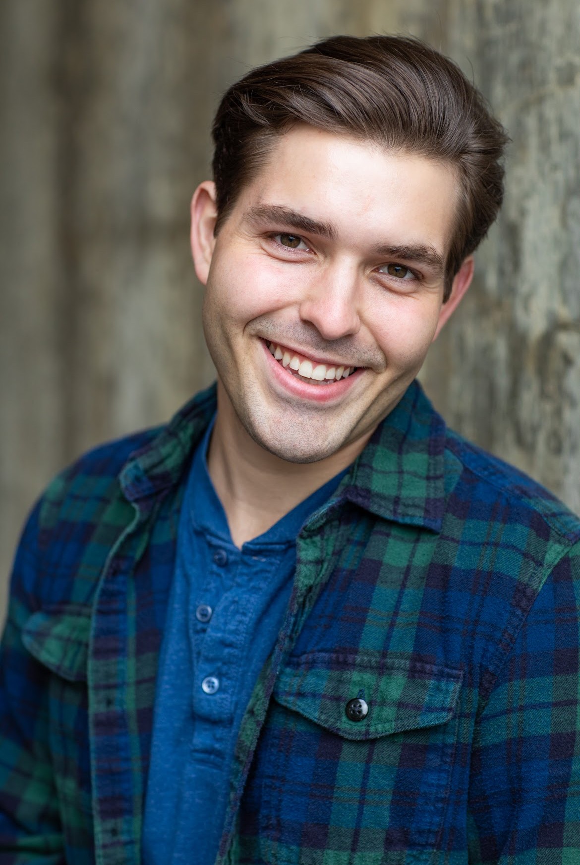 Young man with dark hair smiling, wearing a blue shirt and plaid jacket, standing outdoors near a tree trunk.
