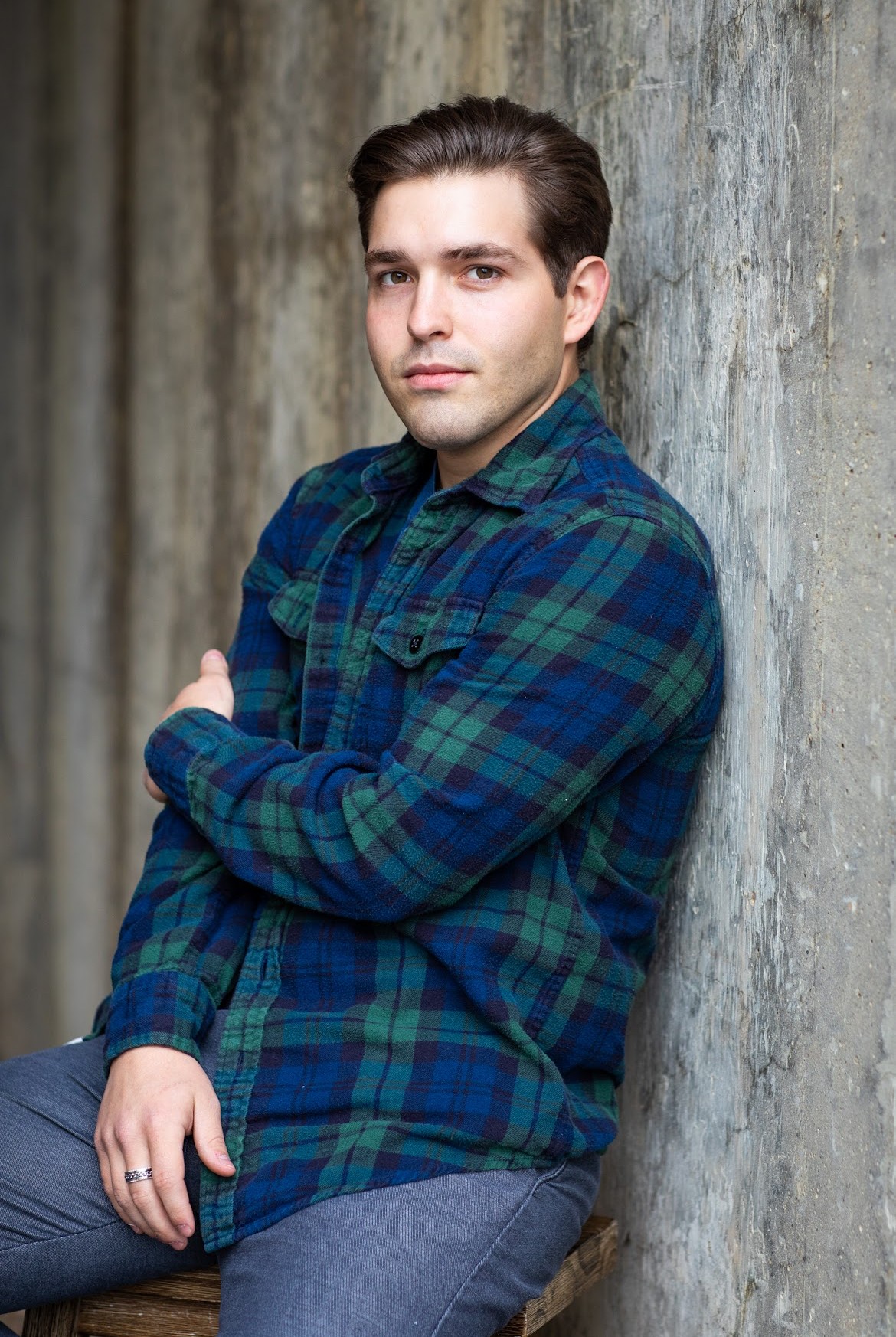 Young man leaning against a concrete wall, wearing a plaid shirt and jeans, with arms crossed.