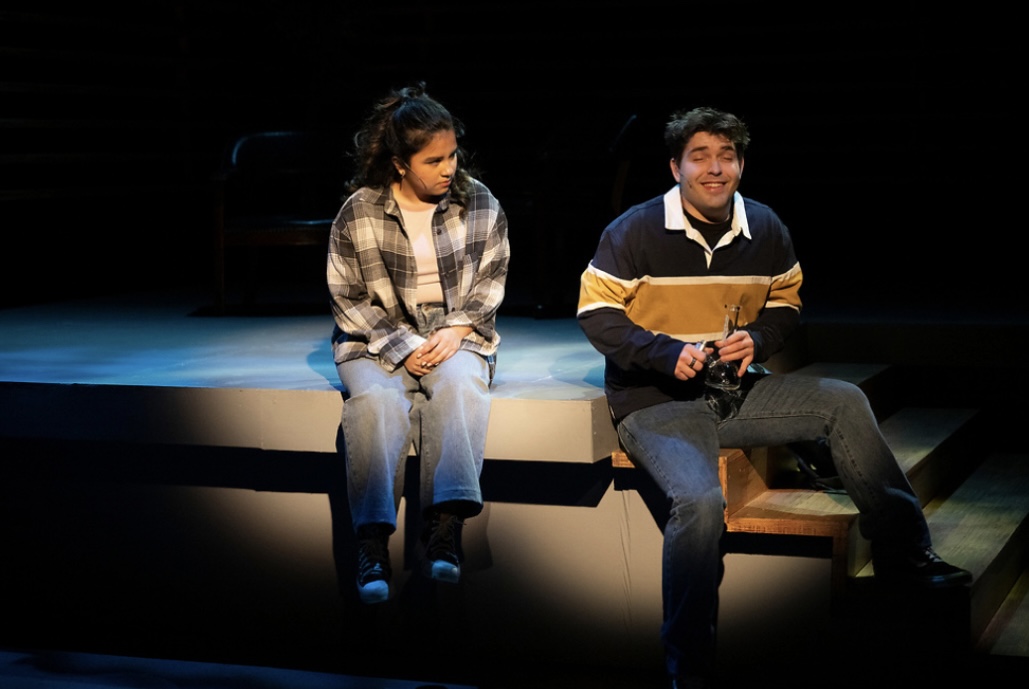 Two people sitting on a stage, one holding a script, with a dark background and spotlight.