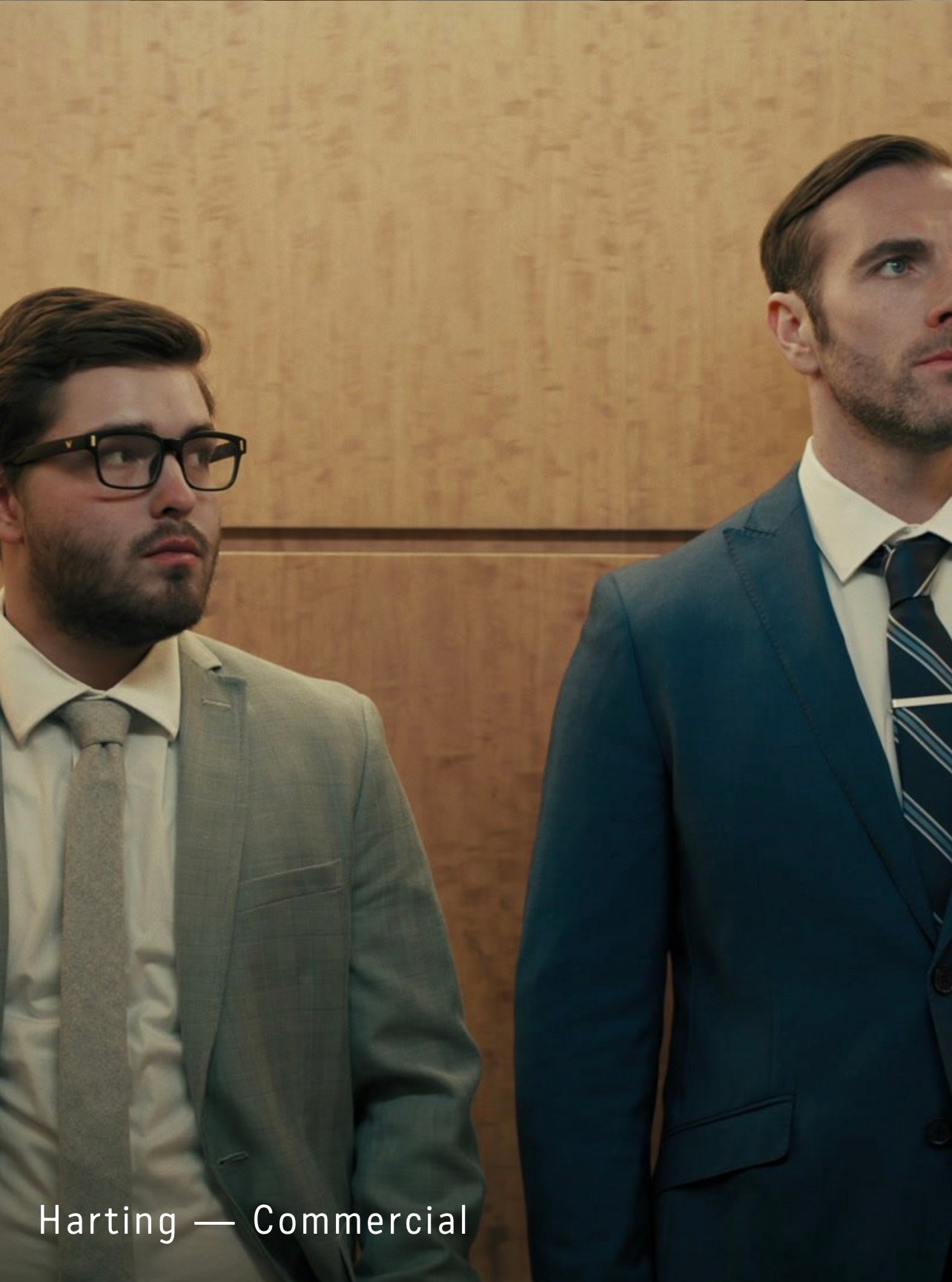 Two men in suits standing indoors against a wooden wall, one with glasses and a beard, the other clean-shaven.