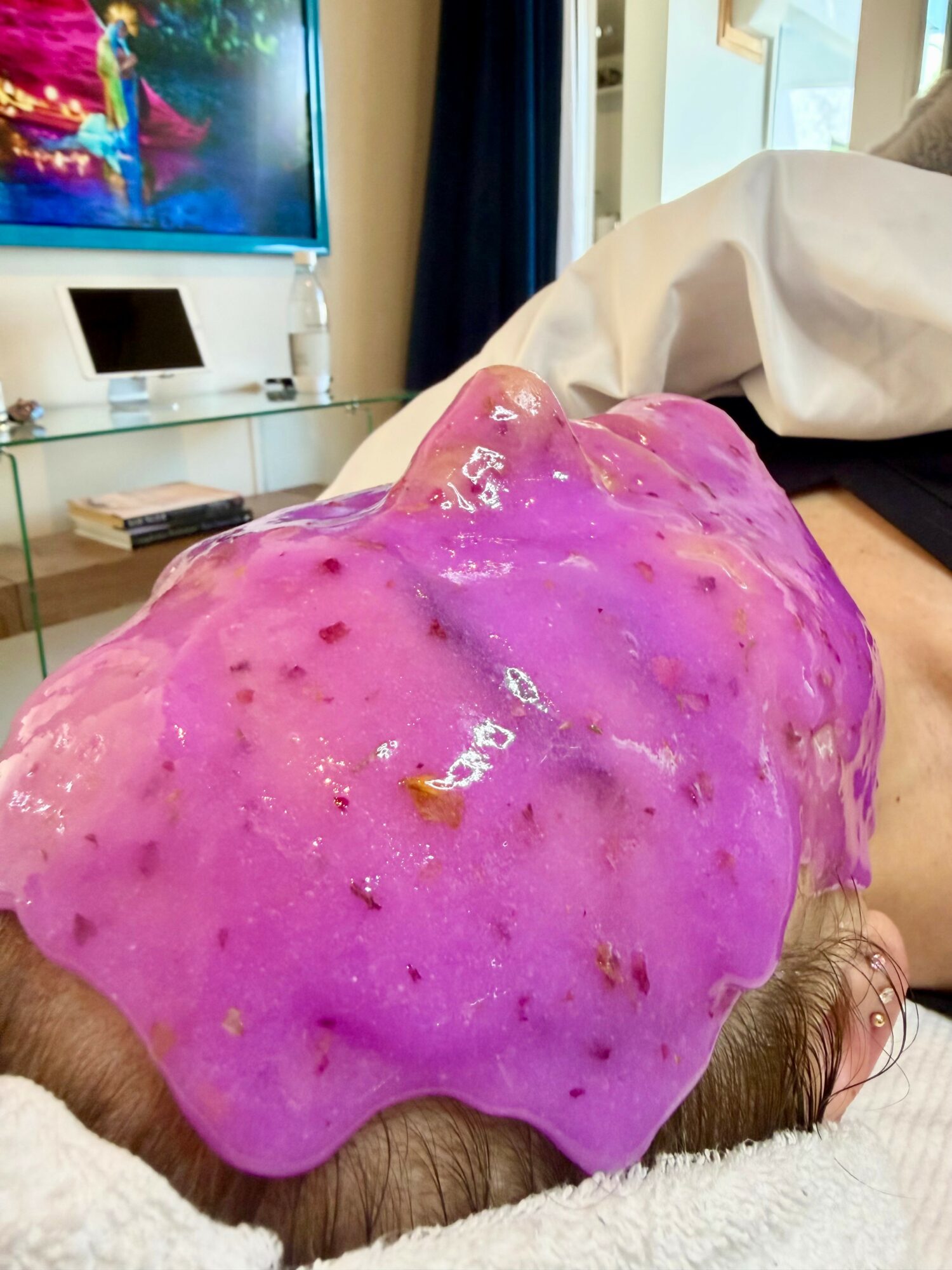 Person lying down with a purple, slimy substance on their head in a room with a TV and glass table.