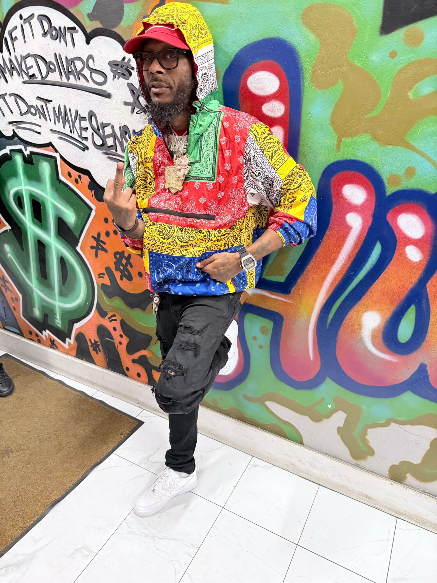 Person wearing colorful clothing and sunglasses standing in front of graffiti wall with dollar sign and text.