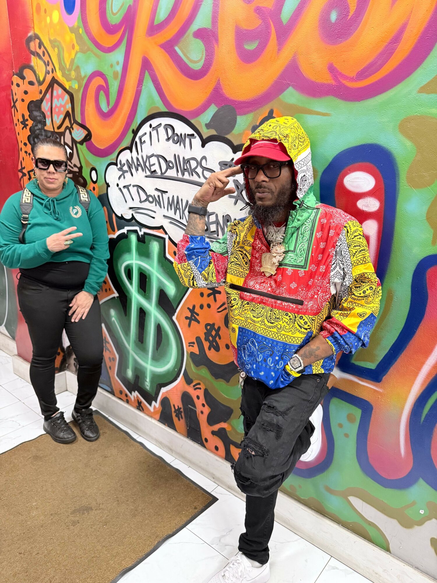 Two people standing in front of a colorful graffiti wall, one with sunglasses and a teal jacket, the other with a red cap and patterned shirt.