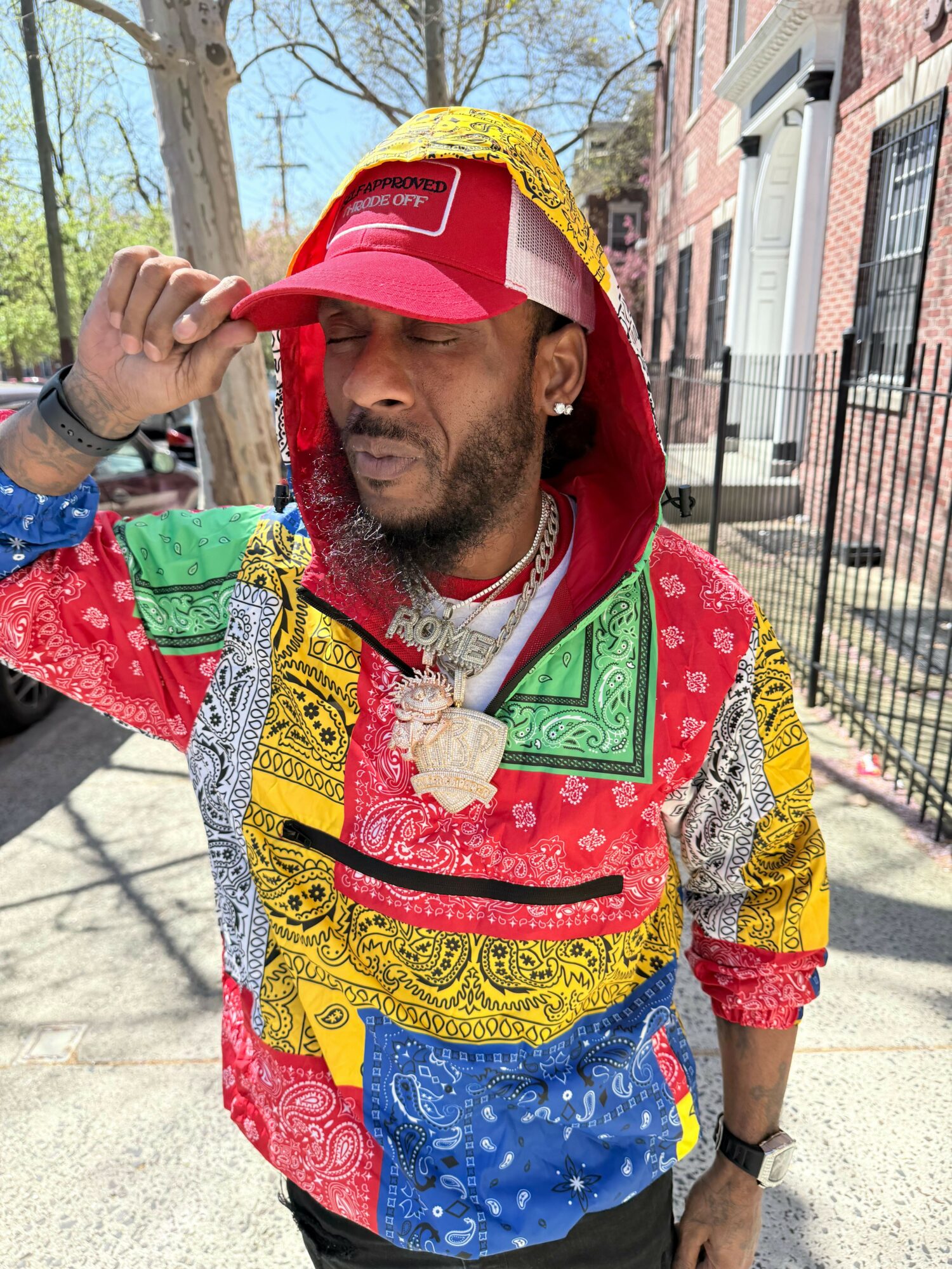 Man wearing colorful patchwork hoodie and red cap, adjusting cap with hand, outdoors on sidewalk with trees and buildings.