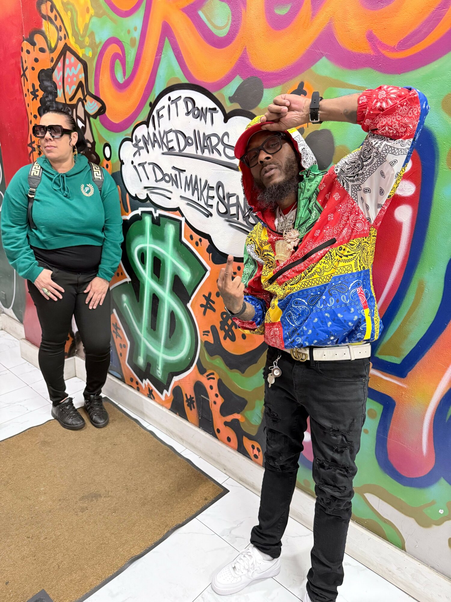 Two people stand in front of a colorful graffiti wall with a dollar sign and speech bubble. One person wears sunglasses and a teal hoodie. The other wears a multicolored jacket and hat.