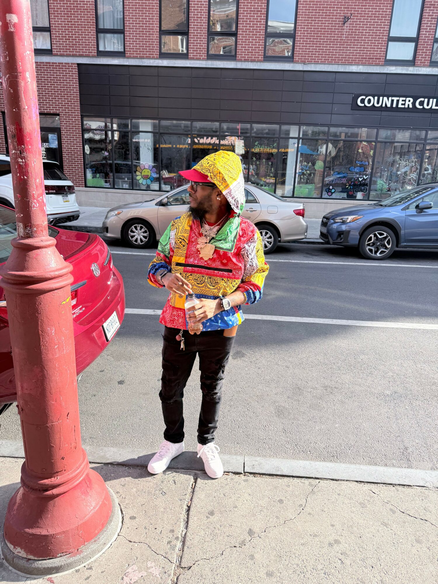 Person standing on sidewalk near a red pole, wearing colorful clothing and a yellow hat, holding a book or tablet.