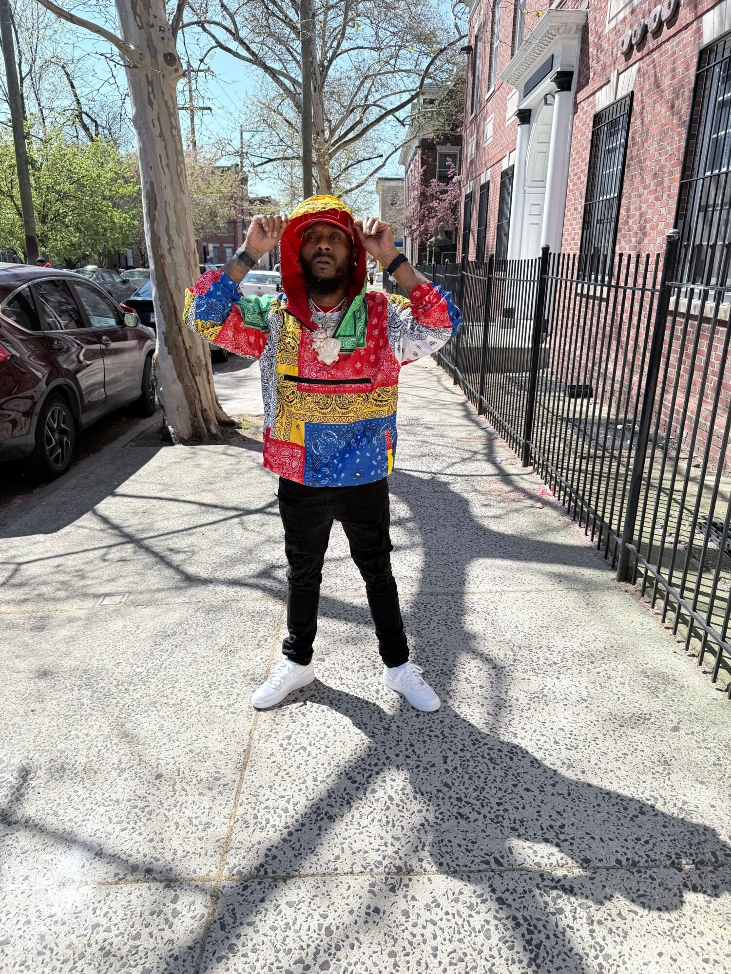 Person dressed in colorful costume standing on sidewalk with trees and buildings in background.