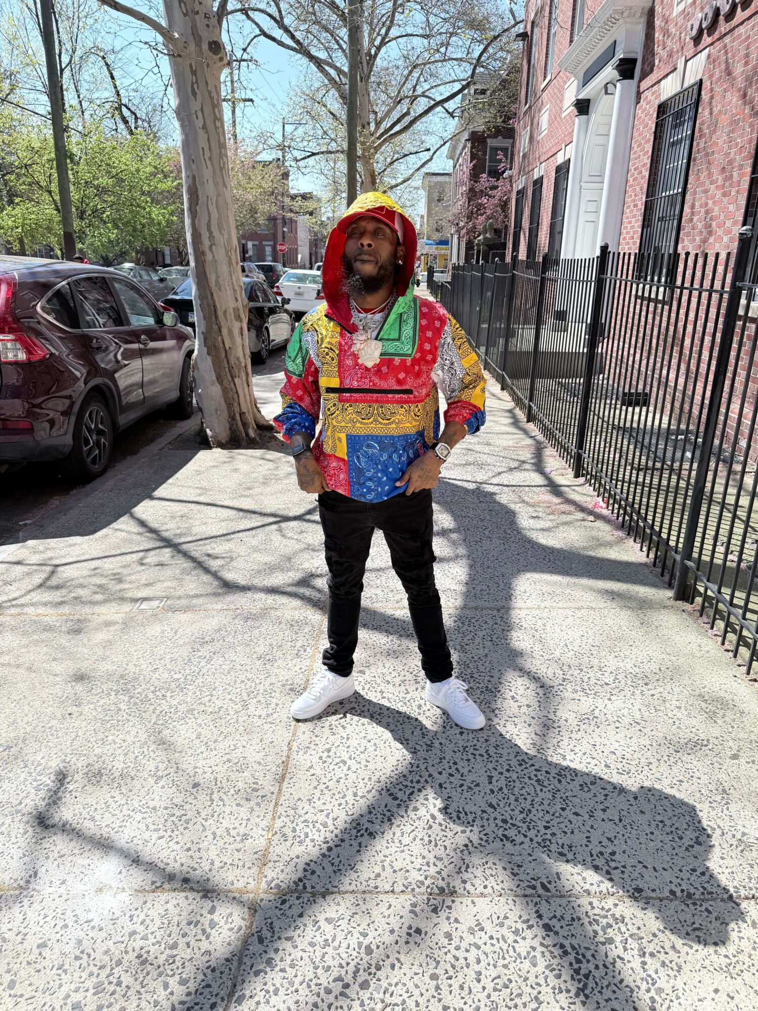 Person standing on sidewalk wearing colorful jacket, hat, and sunglasses, with trees and buildings in background.