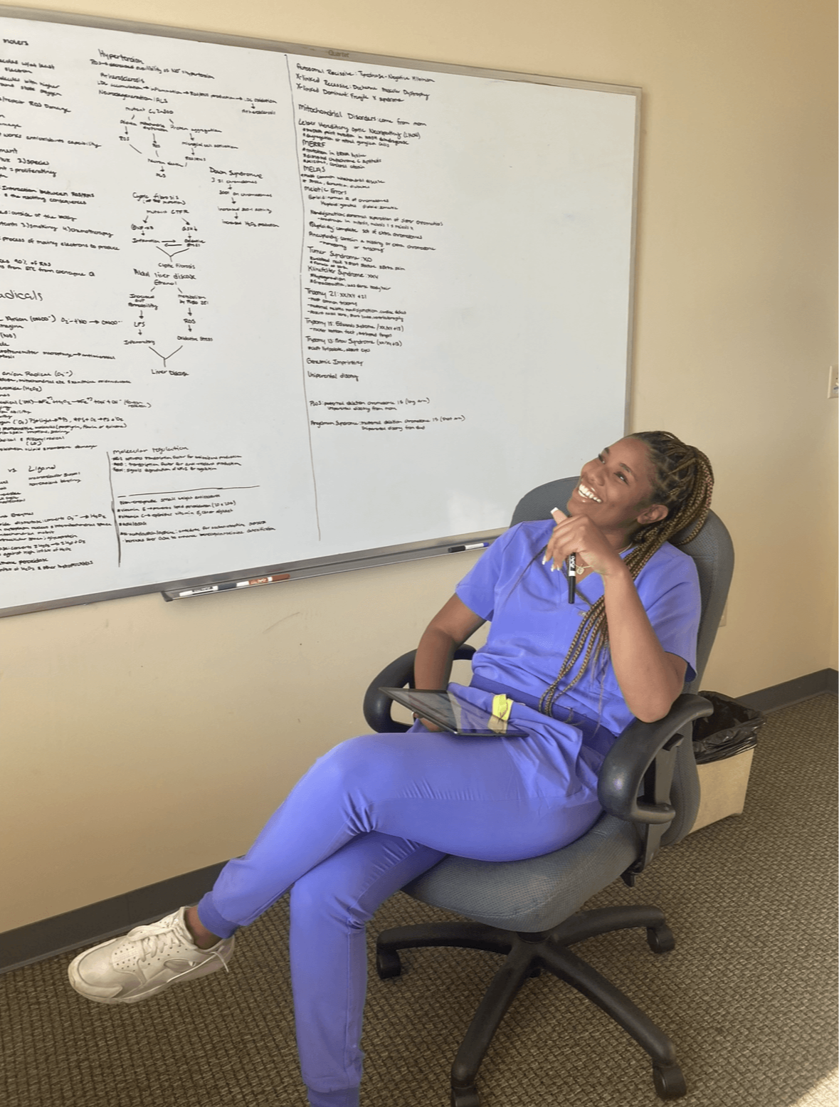 Woman in blue scrubs sitting in chair, resting head on hand, in front of whiteboard with writing.