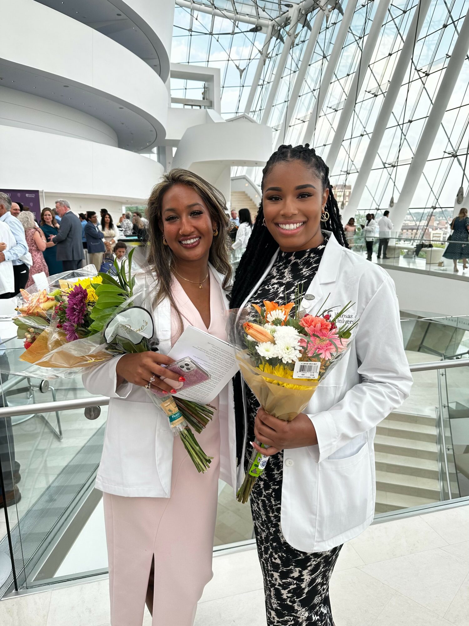 Two women in white coats holding bouquets of flowers, smiling inside a modern building with glass architecture.