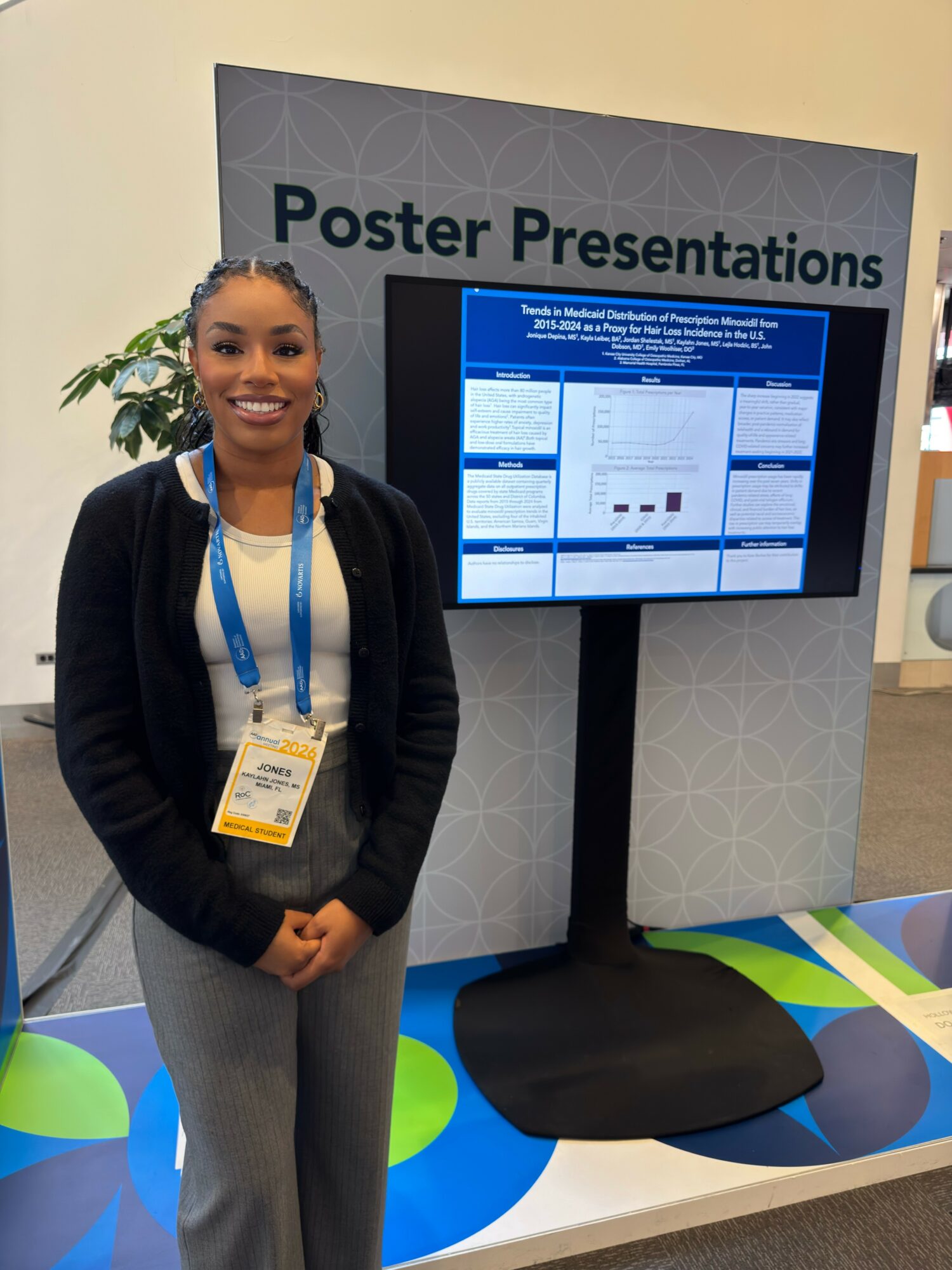 Young woman standing in front of a poster presentation display with a screen showing a presentation slide, smiling.