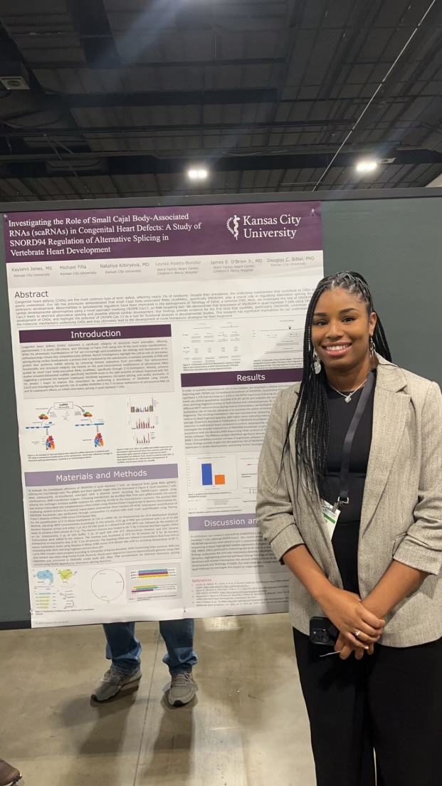 Woman standing in front of research poster at an indoor event, smiling, wearing a beige blazer and black pants.