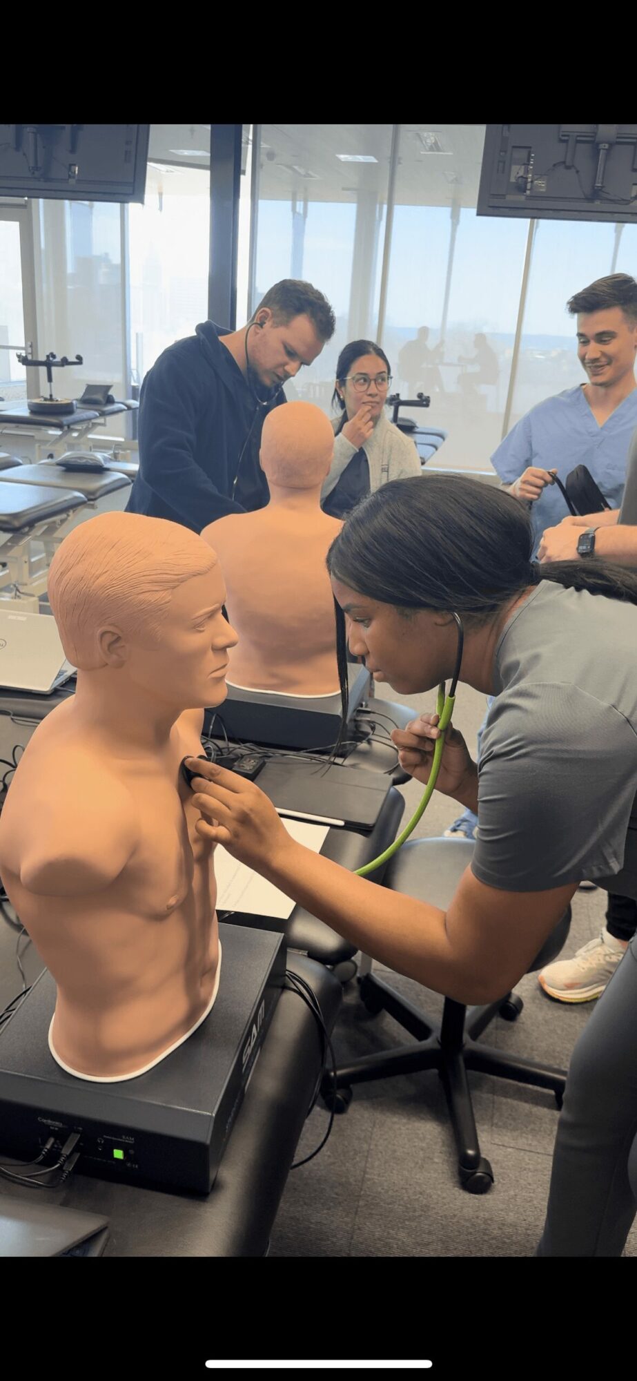 Person practicing auscultation on a medical mannequin in a classroom with three people observing.