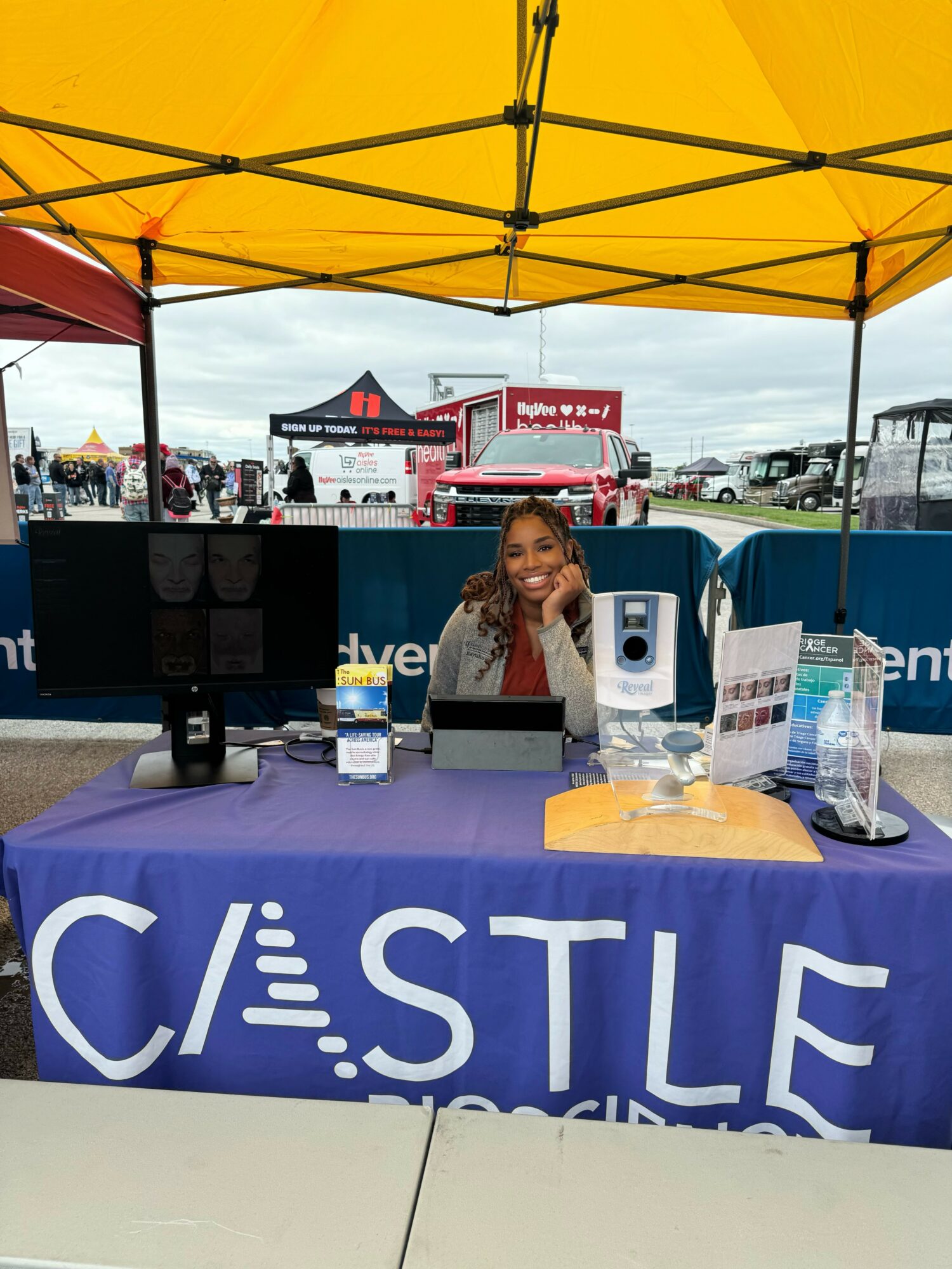 Girl sitting at a booth with a purple tablecloth under a yellow canopy, with trucks and tents in the background.
