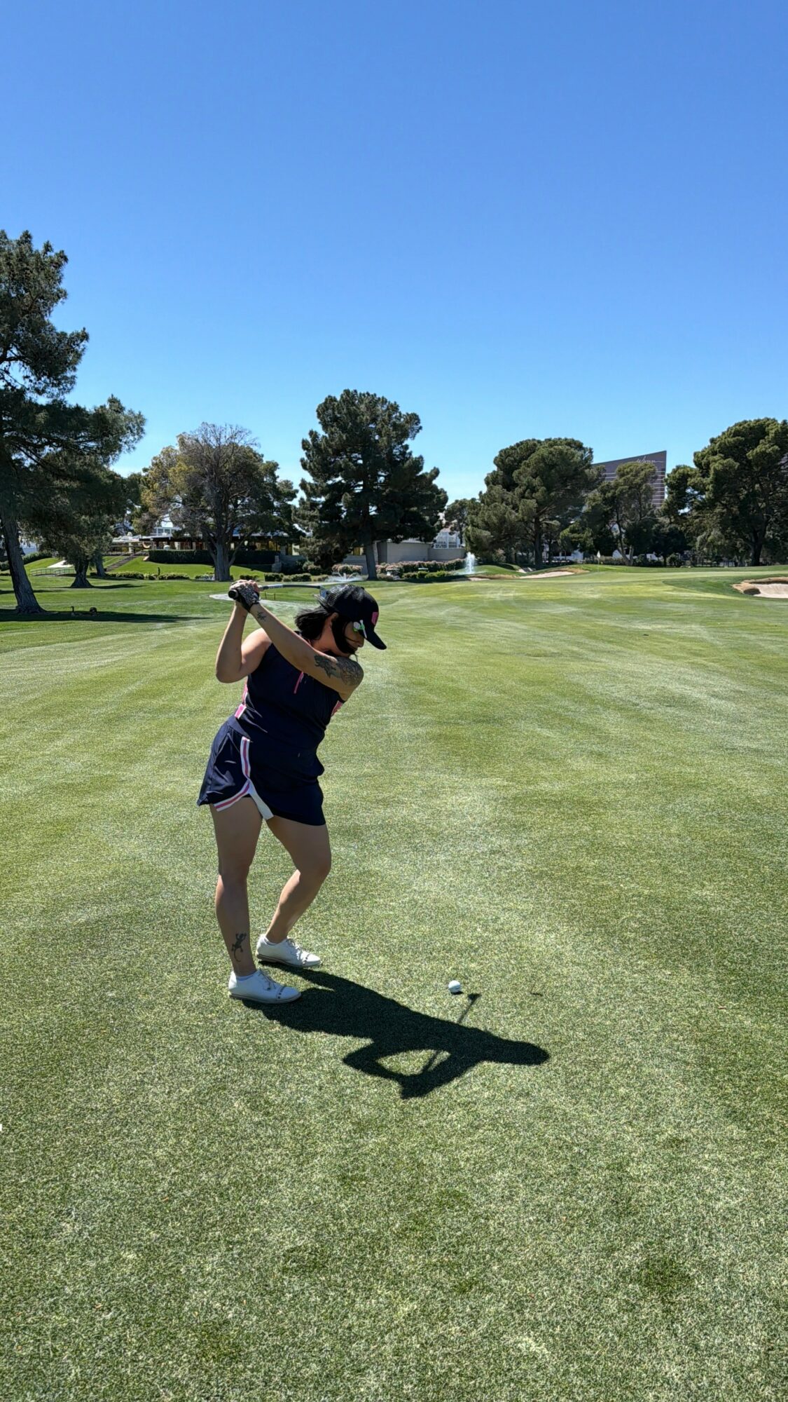 Person swinging golf club on a golf course with trees and blue sky in background.