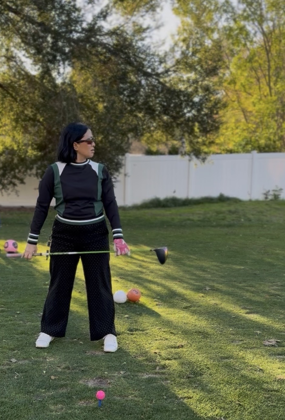 Woman in black outfit with a golf club, standing on grass with trees and a white fence in background.