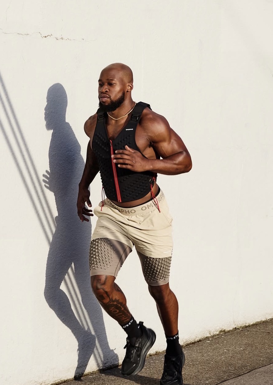 Muscular man running outdoors against a white wall, shadow cast on wall, wearing shorts, tank top, and sneakers.