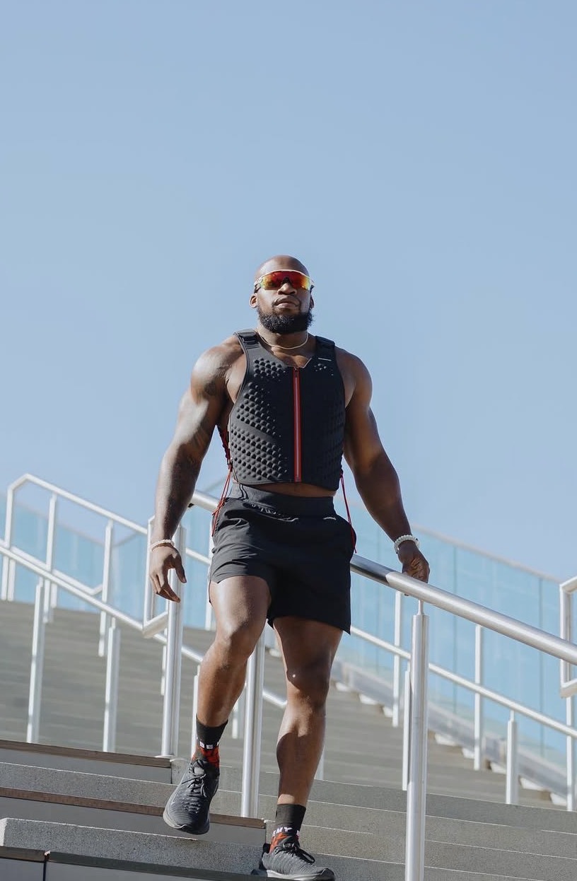 Athletic man in sportswear walking down outdoor stairs with railing, wearing sunglasses and a hydration vest, against a clear blue sky.