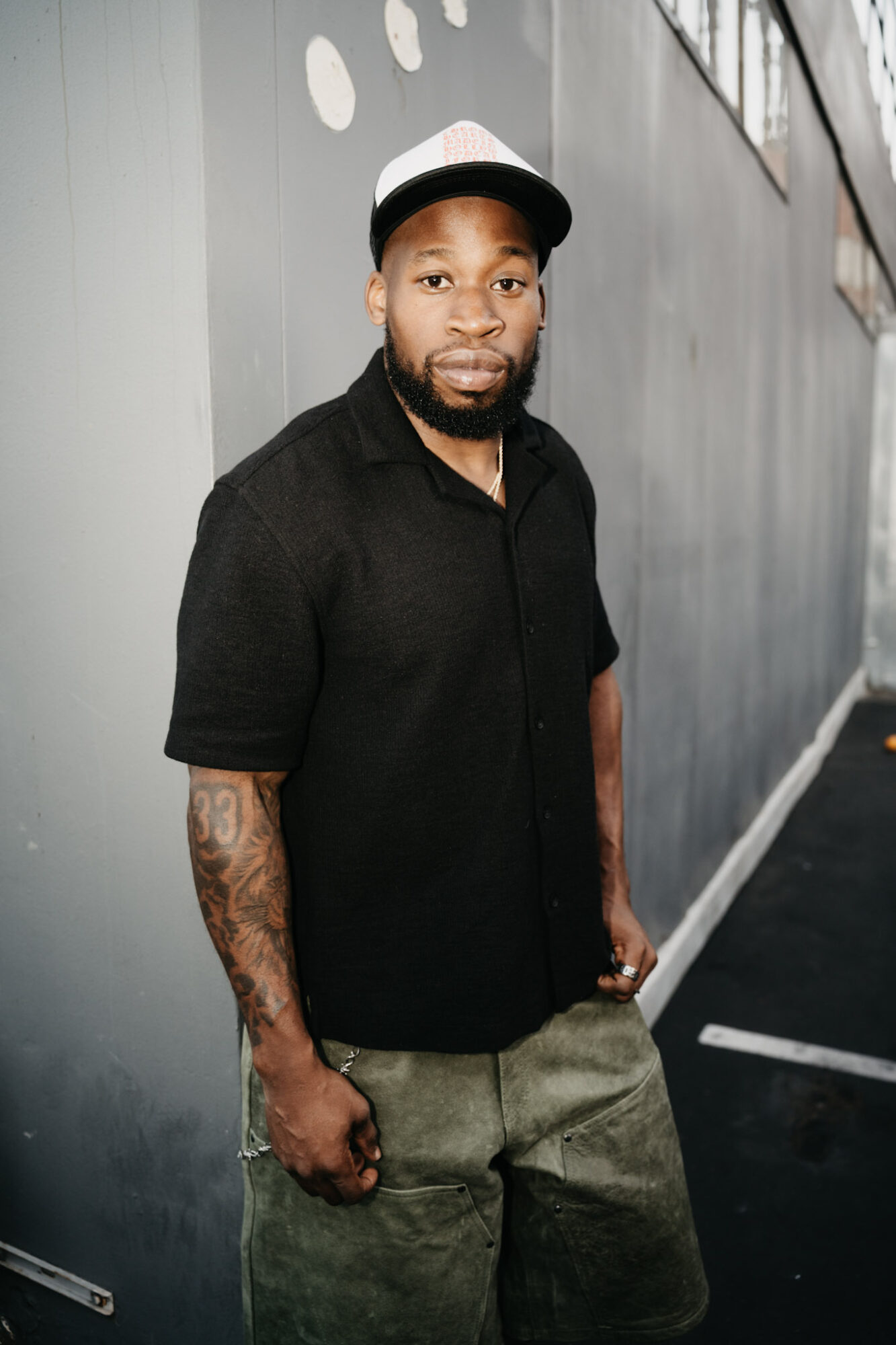 Young man with a beard, wearing a black shirt and a white cap, standing against a gray wall.