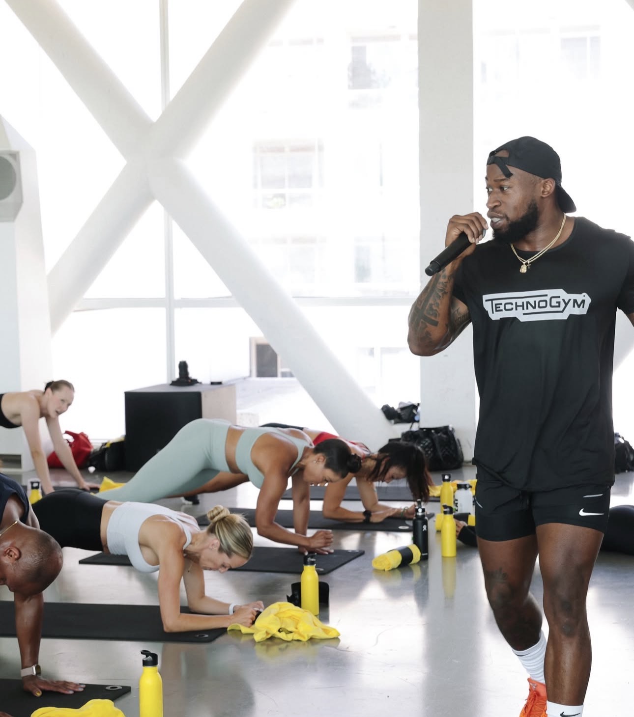 Instructor speaking into microphone during fitness class with participants doing plank exercises on mats.