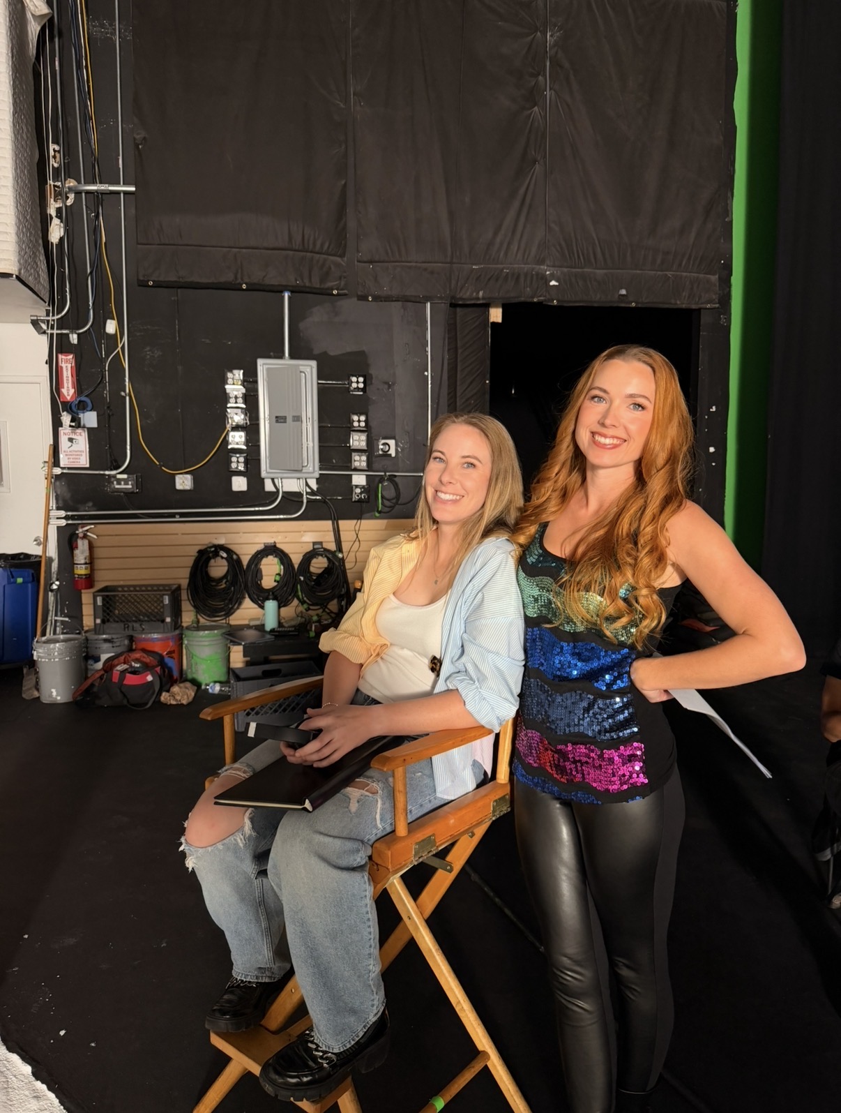 Two women smiling, one sitting on a director's chair, the other standing beside her, in a studio setting with equipment behind them.