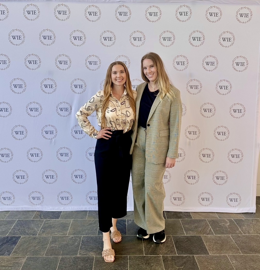 Two women standing together in front of a backdrop with repeated logo pattern, smiling, one with hand on hip, the other with arm around shoulder.