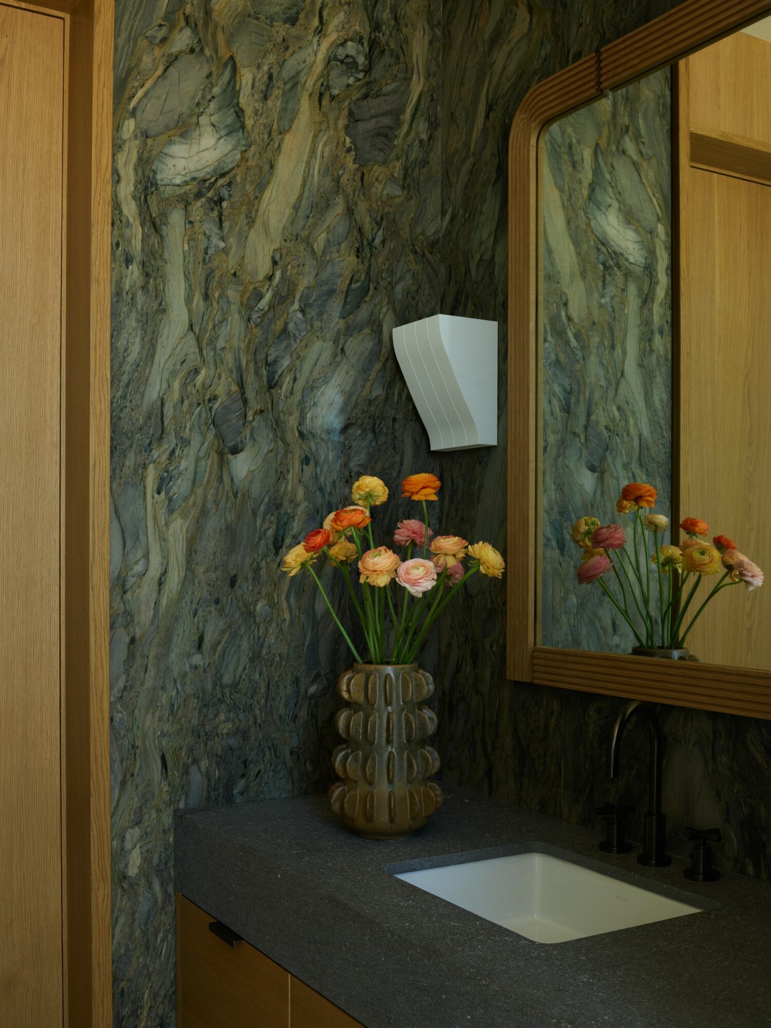 Bathroom with a dark countertop, a vase with colorful flowers, a mirror, and a wall-mounted light fixture.