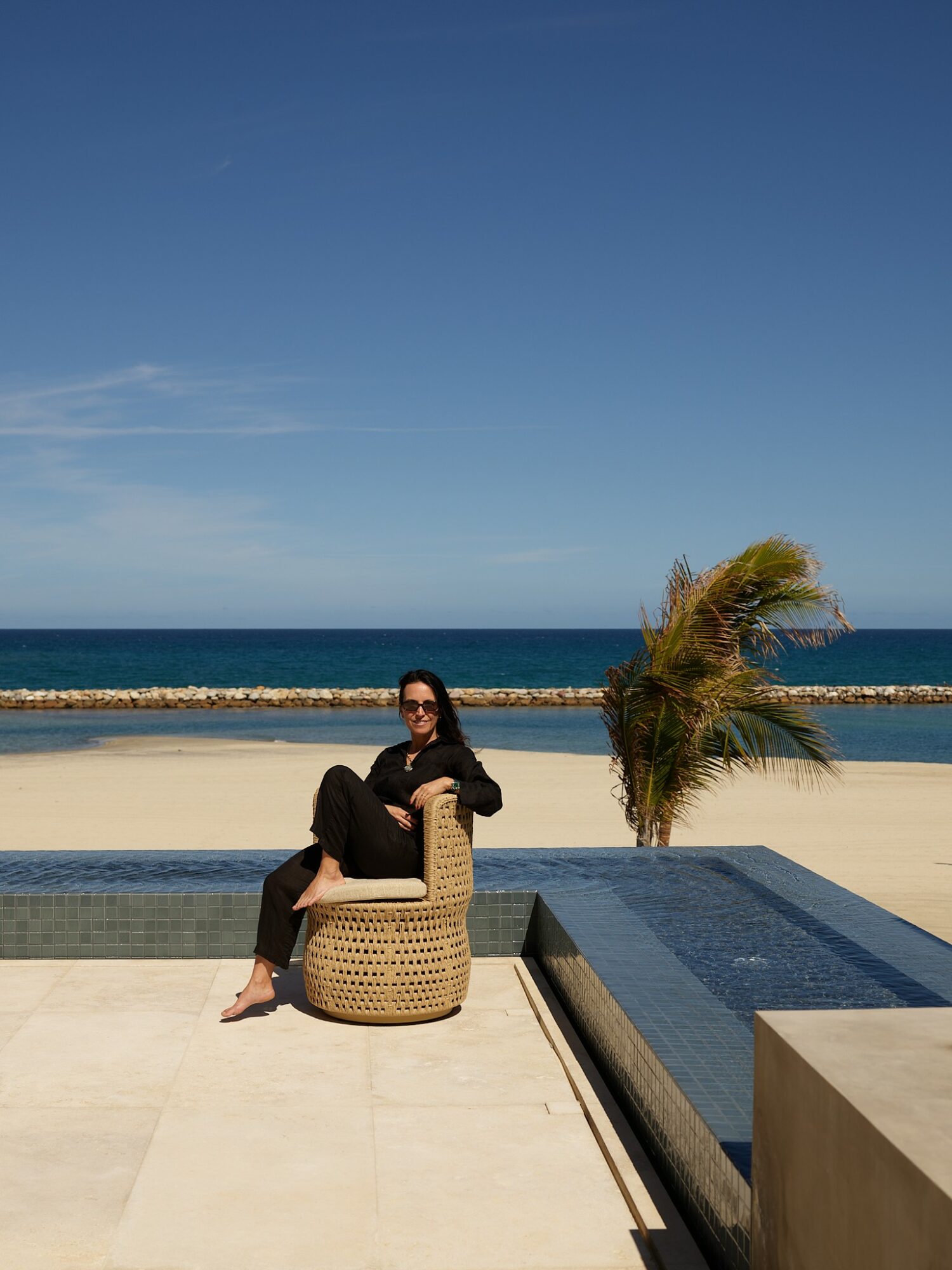 Person sitting on a wicker chair near a pool with a palm tree and ocean background.