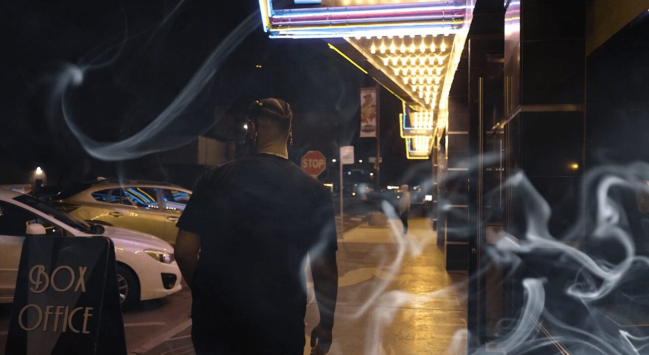 Person walking on a city sidewalk at night with illuminated signs and cars parked nearby, smoke or mist in the foreground.