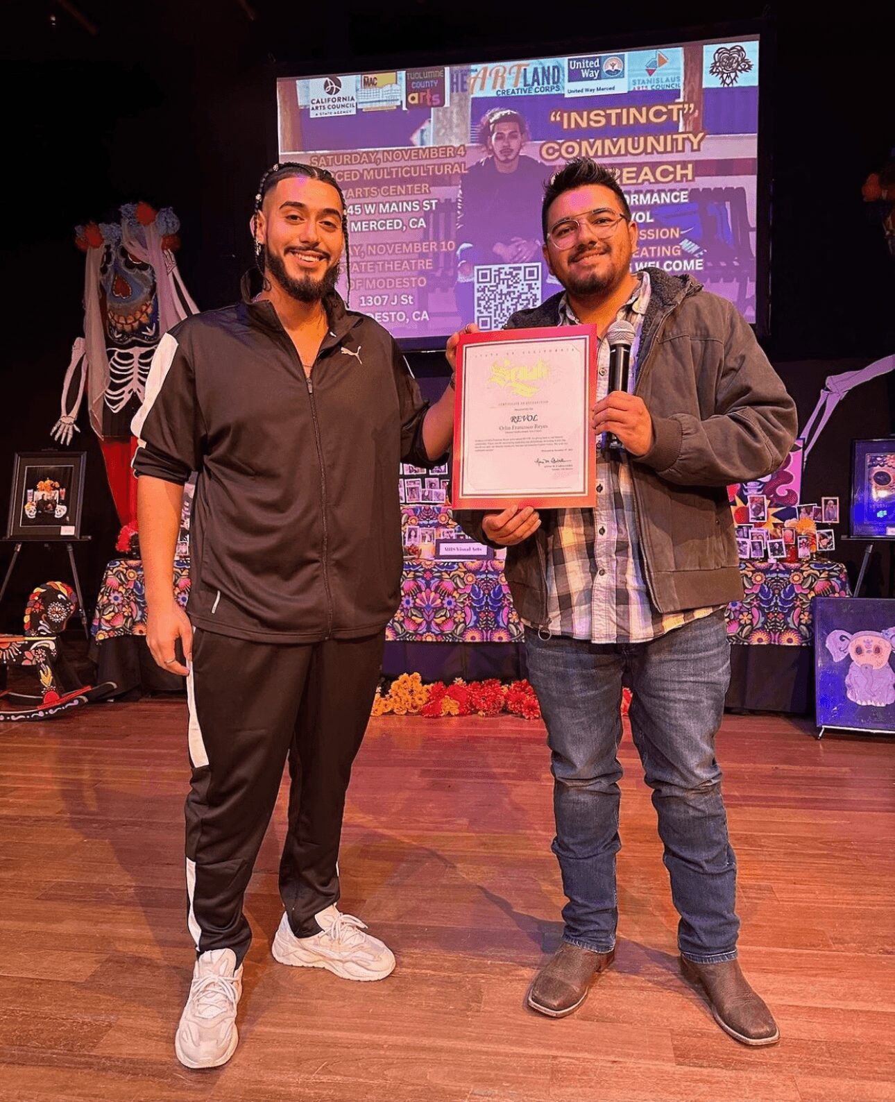 Two men stand on stage, one holding a framed certificate, smiling. Background features a colorful screen and decorations.