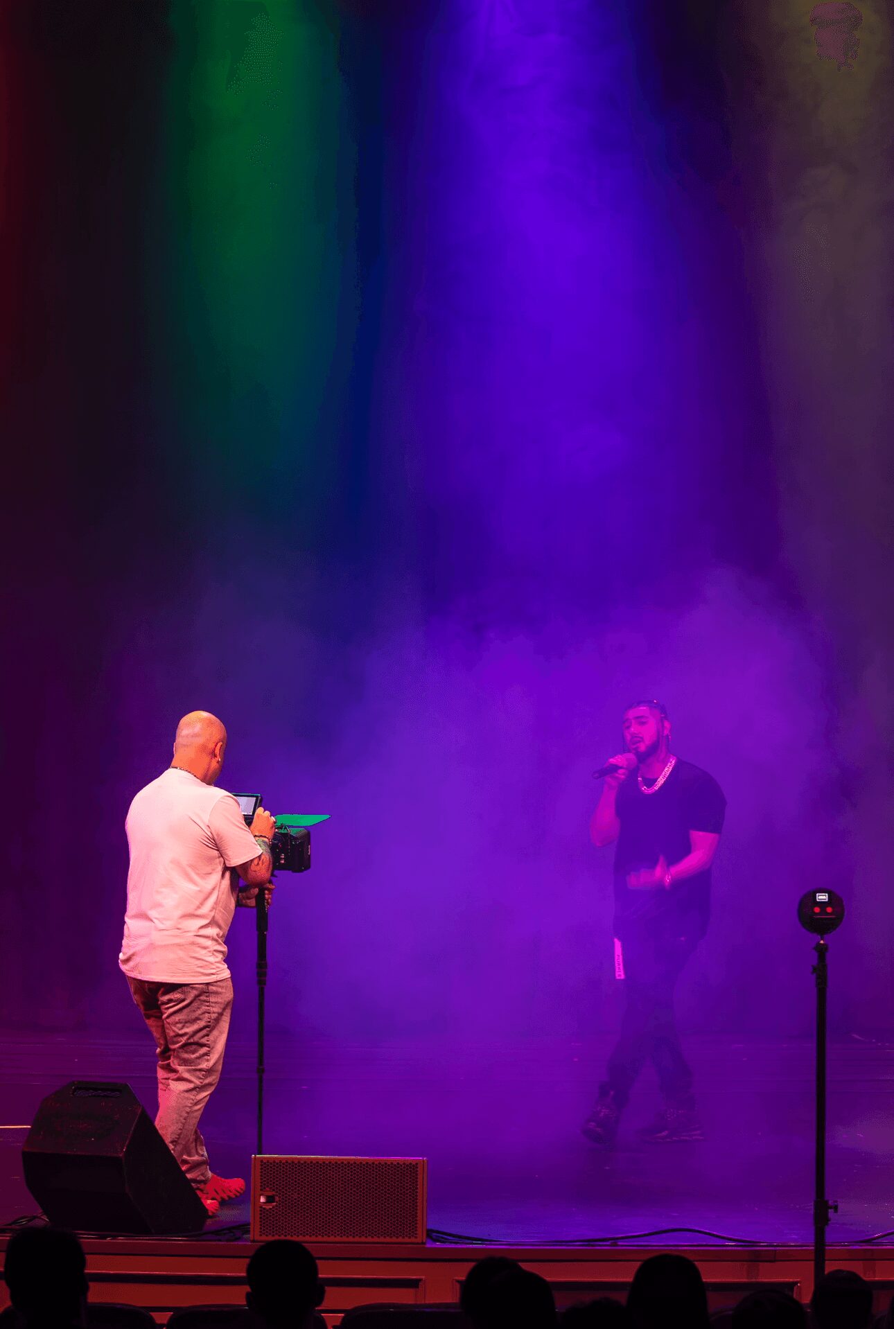 Two people on stage with colorful lighting and fog, one holding a camera and the other singing into a microphone.
