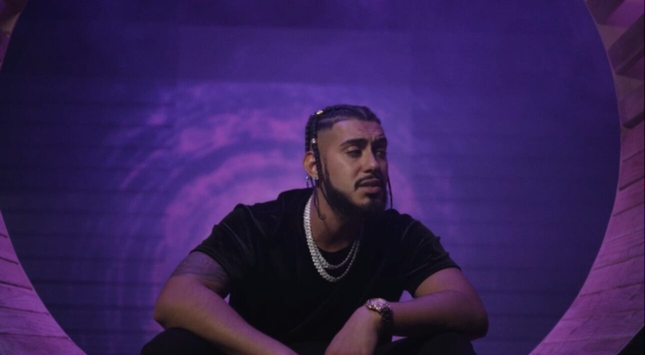 Man with beard and braided hair, wearing black shirt and jewelry, sitting against purple background.