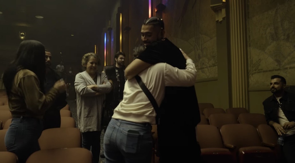 Person hugging another person in a dimly lit room with chairs and other people in the background.