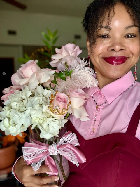 Woman smiling holding a bouquet of pink and white flowers with a pink ribbon, wearing a pink shirt and maroon apron.